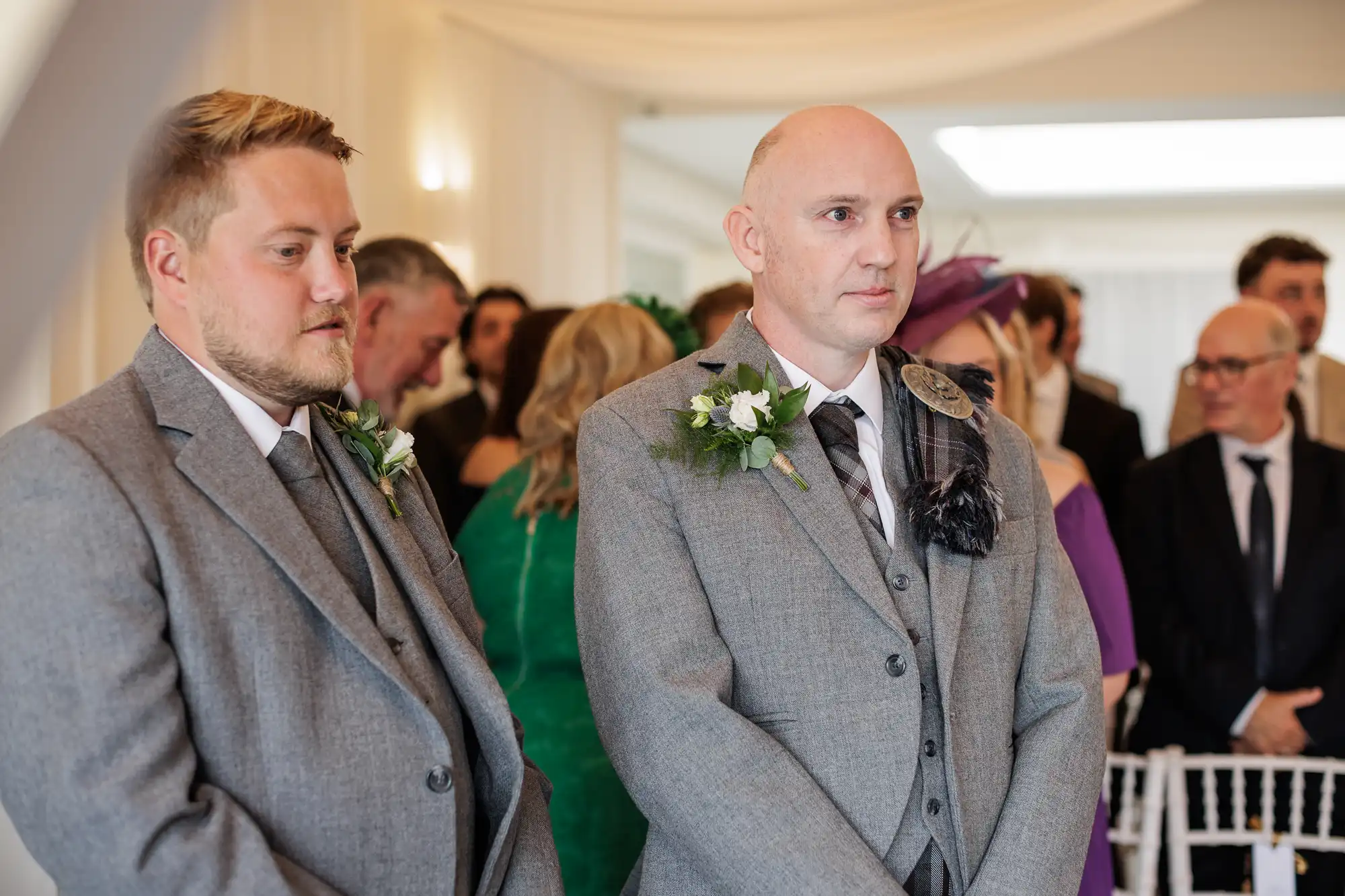 Two men in grey suits with buttonholes stand side by side at a formal event, possibly a wedding. One is younger with blond hair and beard, the other bald. People dressed in formal attire are visible in the blurred background.