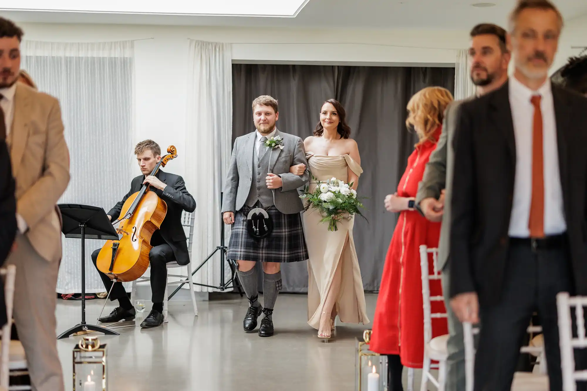 A man in a kilt and a woman in a beige dress holding white flowers walk arm-in-arm down an aisle. Guests stand on either side. A cellist plays music to the left. The bright room has white walls, curtains, and modern decor.