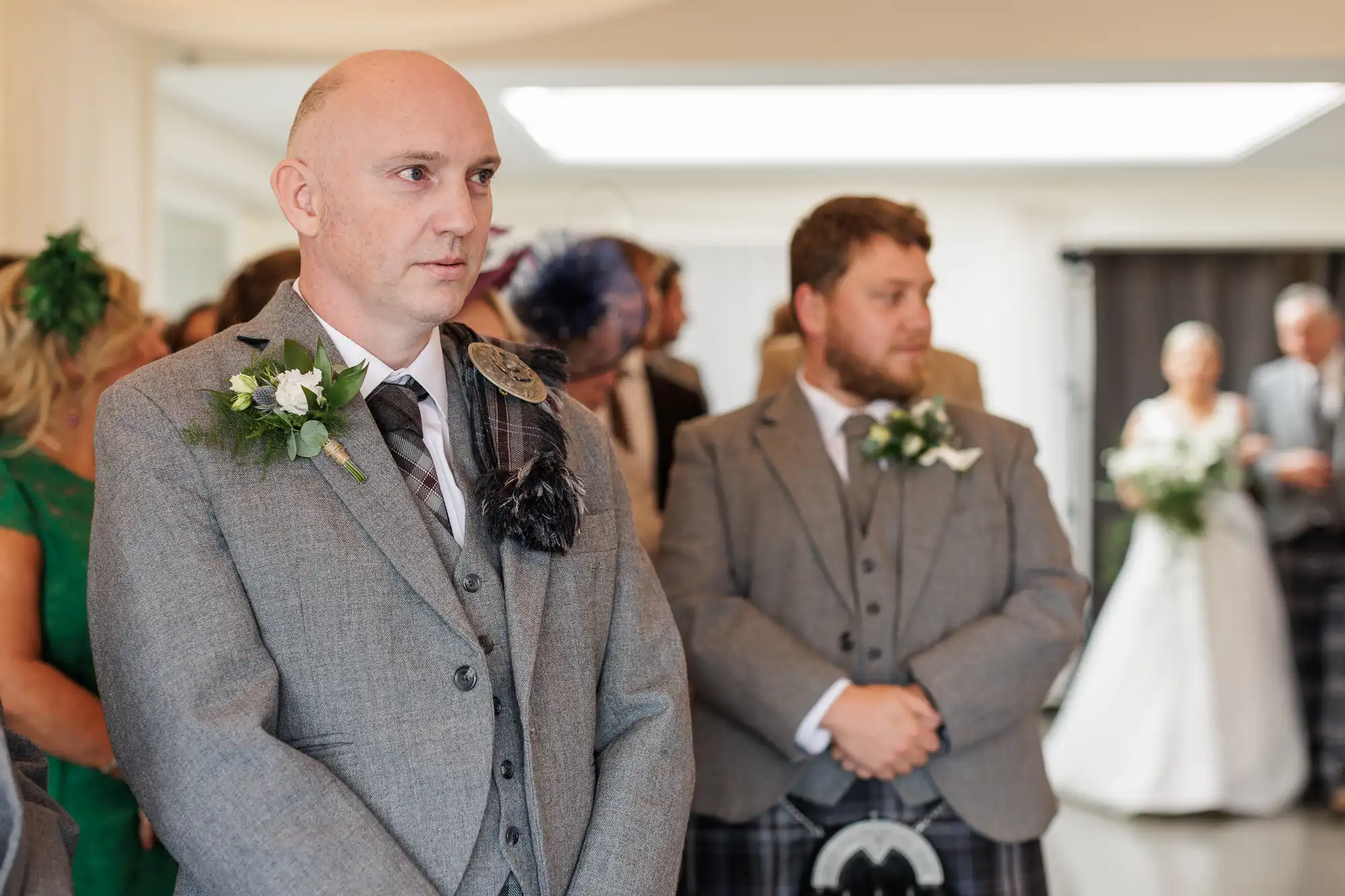 Two men in grey suits with buttonholes stand indoors at a wedding ceremony. The man in front is bald and looks serious; the other has brown hair and beard. People and a bride in a white dress are blurred in the background.