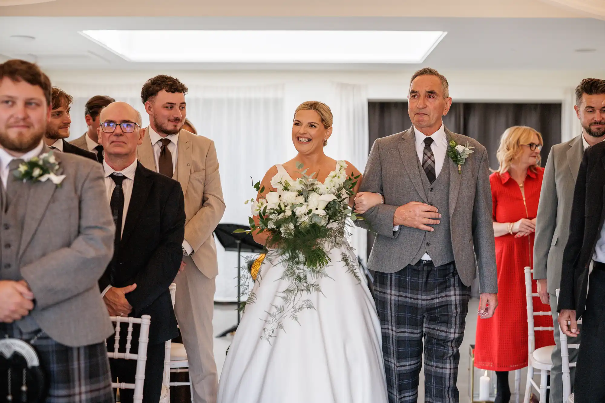 A smiling bride in a white gown holds a bouquet and walks arm-in-arm with an older man in a grey jacket and checked trousers down an aisle, surrounded by guests dressed in formal attire, some standing and watching, inside a bright ceremony room.