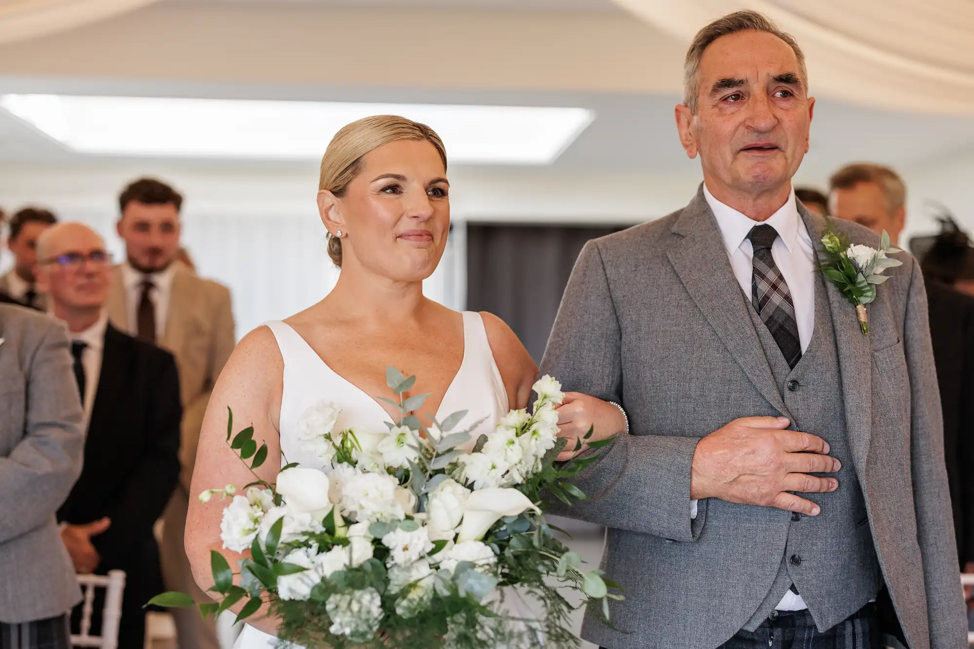 A bride in a white dress holding a bouquet of white flowers walks arm-in-arm with an older man in a grey suit and checked waistcoat. They are indoors, surrounded by formally dressed guests, and both appear emotional and happy.