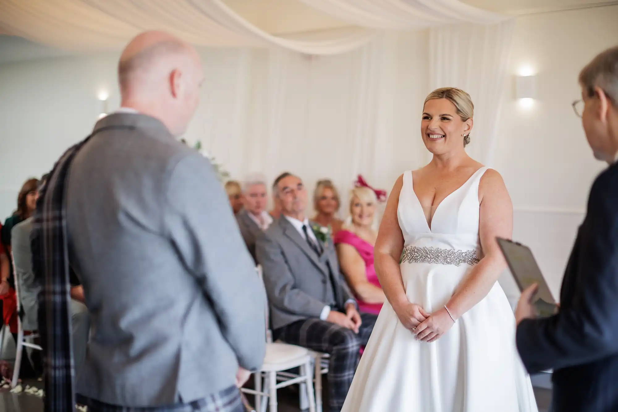 A smiling bride in a white dress stands facing a man in a suit during a wedding ceremony, with seated guests watching in the background. The room is decorated with white drapes and soft lighting, creating an elegant atmosphere.