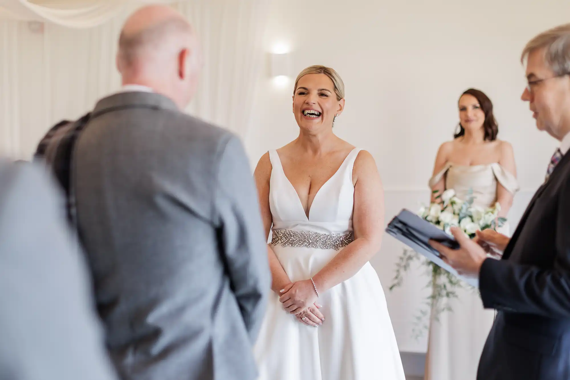 A smiling bride in a white dress with a jewelled belt stands facing a man in a suit. A woman in a pale dress holding flowers and another man reading from a book are nearby. The room is softly lit with white drapes and subtle decorations.