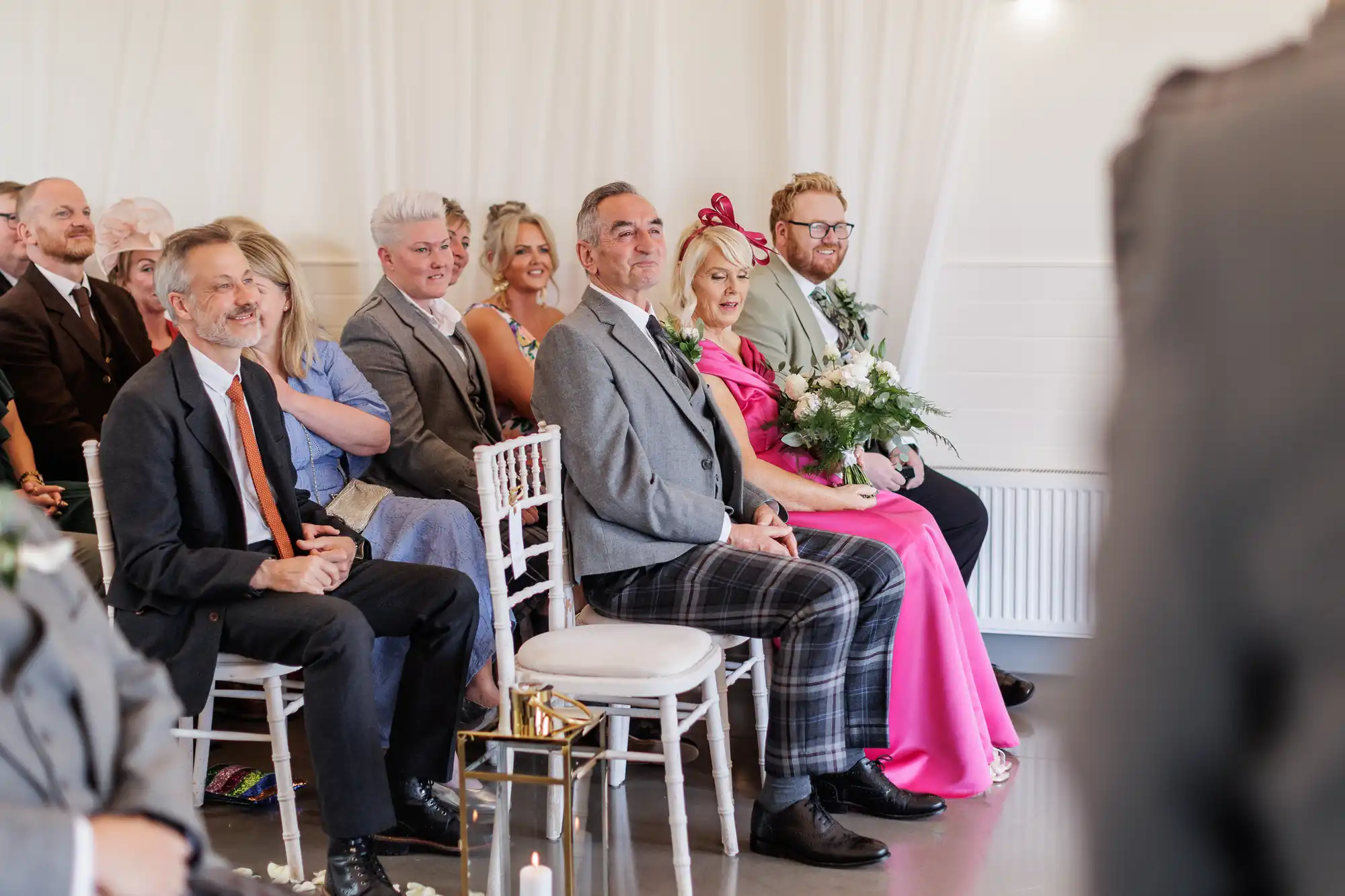 Wedding guests sit on white chairs, smiling and looking ahead. Most are dressed in formal attire; one woman wears a bright pink dress and hat, holding a bouquet. The background has white walls and draped curtains, giving a bright, elegant atmosphere.
