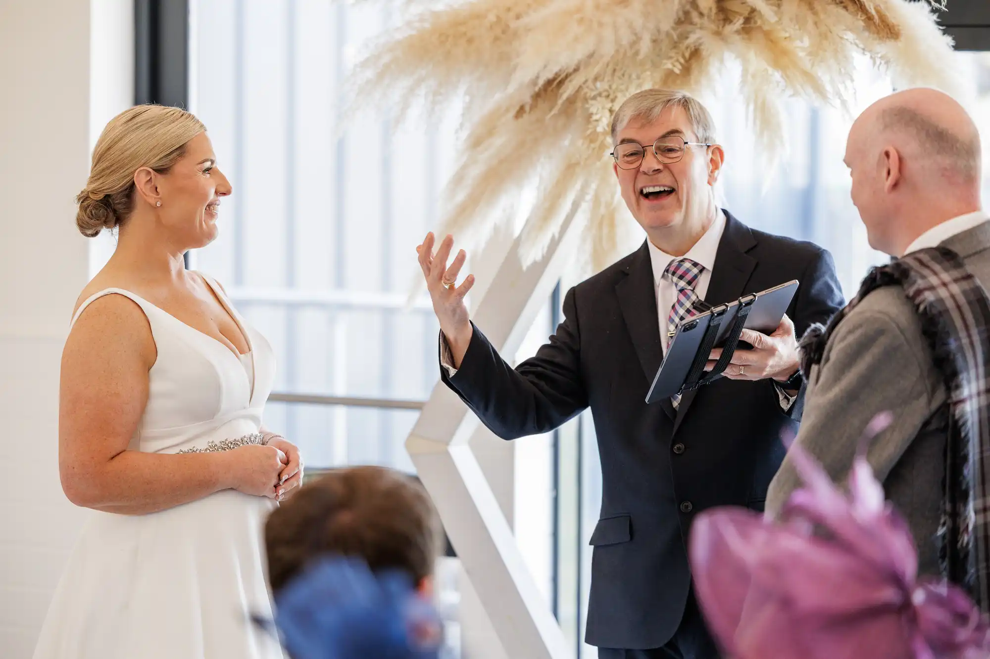 A bride in a white dress smiles at a groom in a grey suit as an officiant in glasses and a suit enthusiastically speaks, holding a tablet. They stand indoors under decorative pampas grass, with guests seated in the foreground.