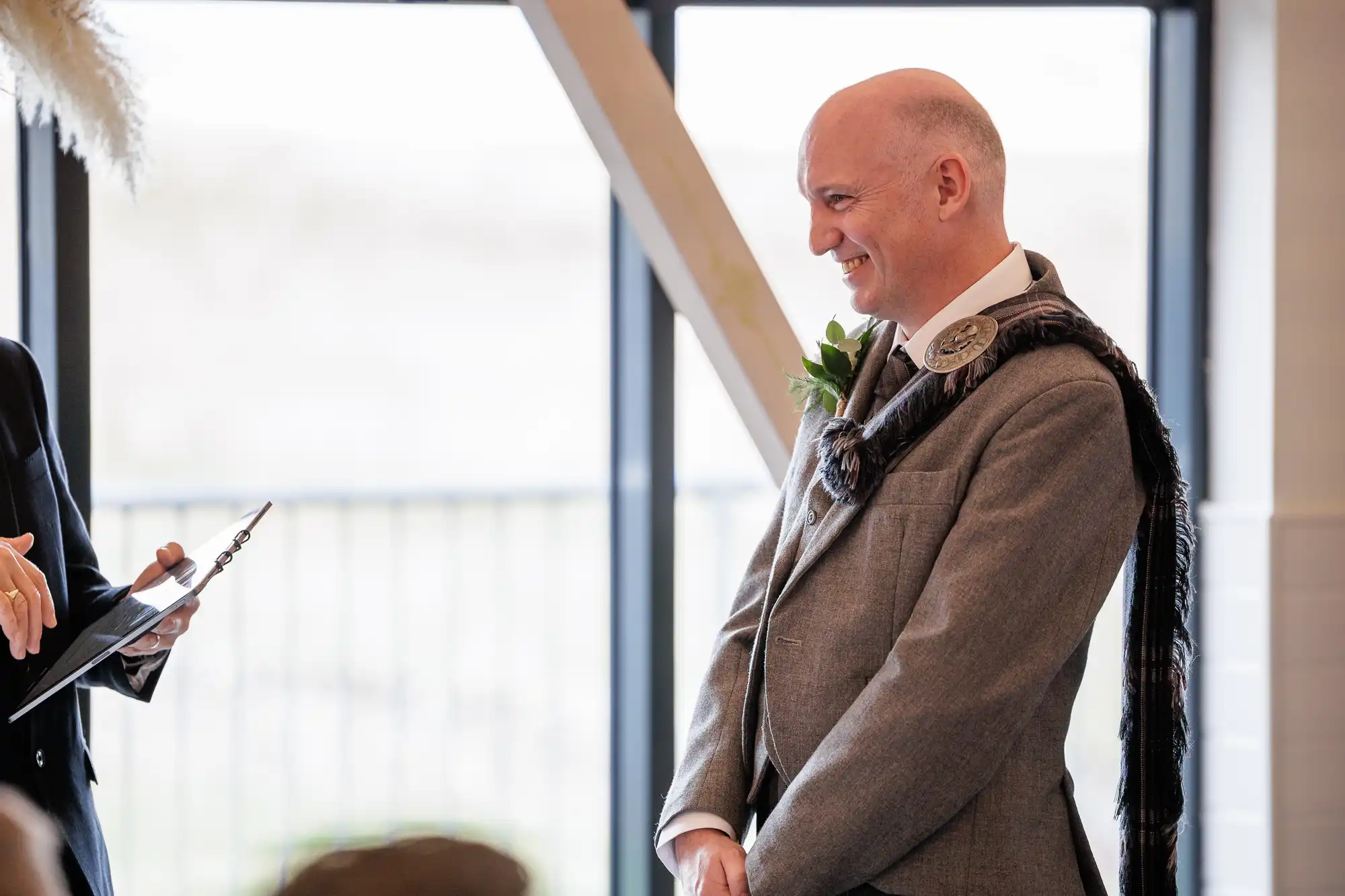 A smiling bald man in a grey suit with a checked sash and large decorative brooch stands indoors, facing a person holding a clipboard. The background features tall windows letting in natural light.