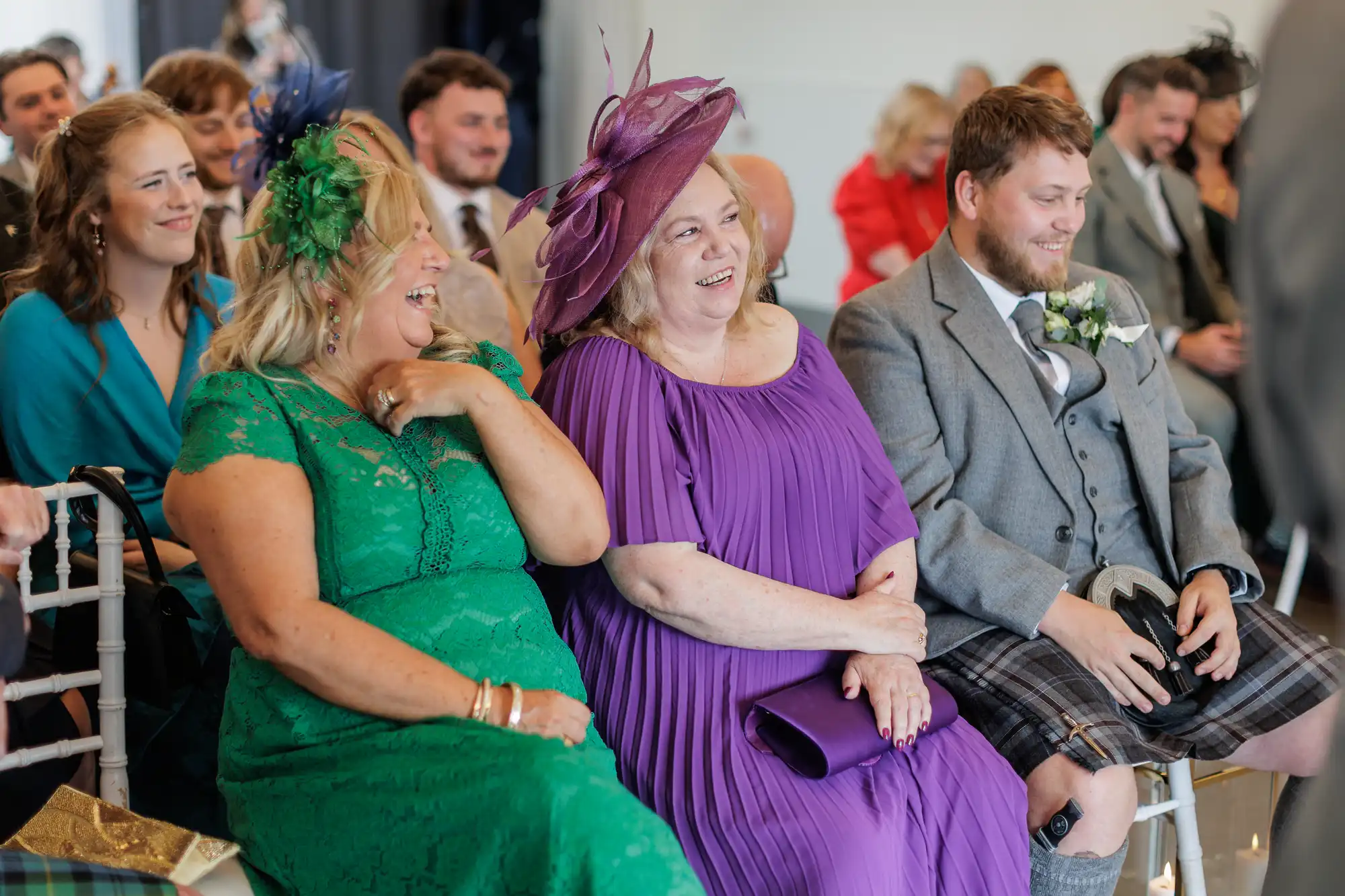 Three people sit and smile in the front row at a wedding ceremony. One woman wears a green dress and hat, another wears a purple dress and matching hat, and a man wears a grey jacket with a kilt. Guests in colourful attire are seated behind them.