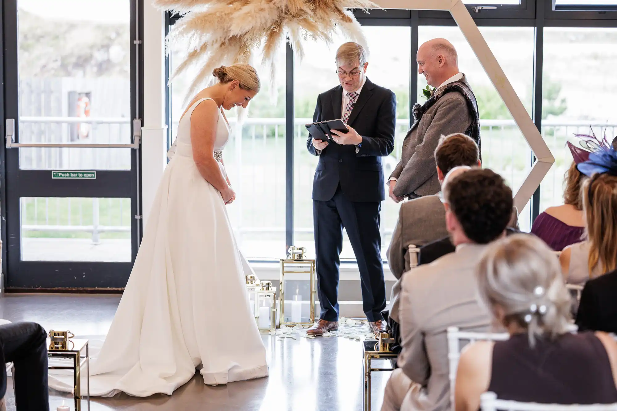 A bride in a white dress and a groom in a dark suit stand before an officiant holding a tablet during an indoor wedding ceremony. Guests are seated, and a floral arrangement with pampas grass decorates the bright backdrop behind them.