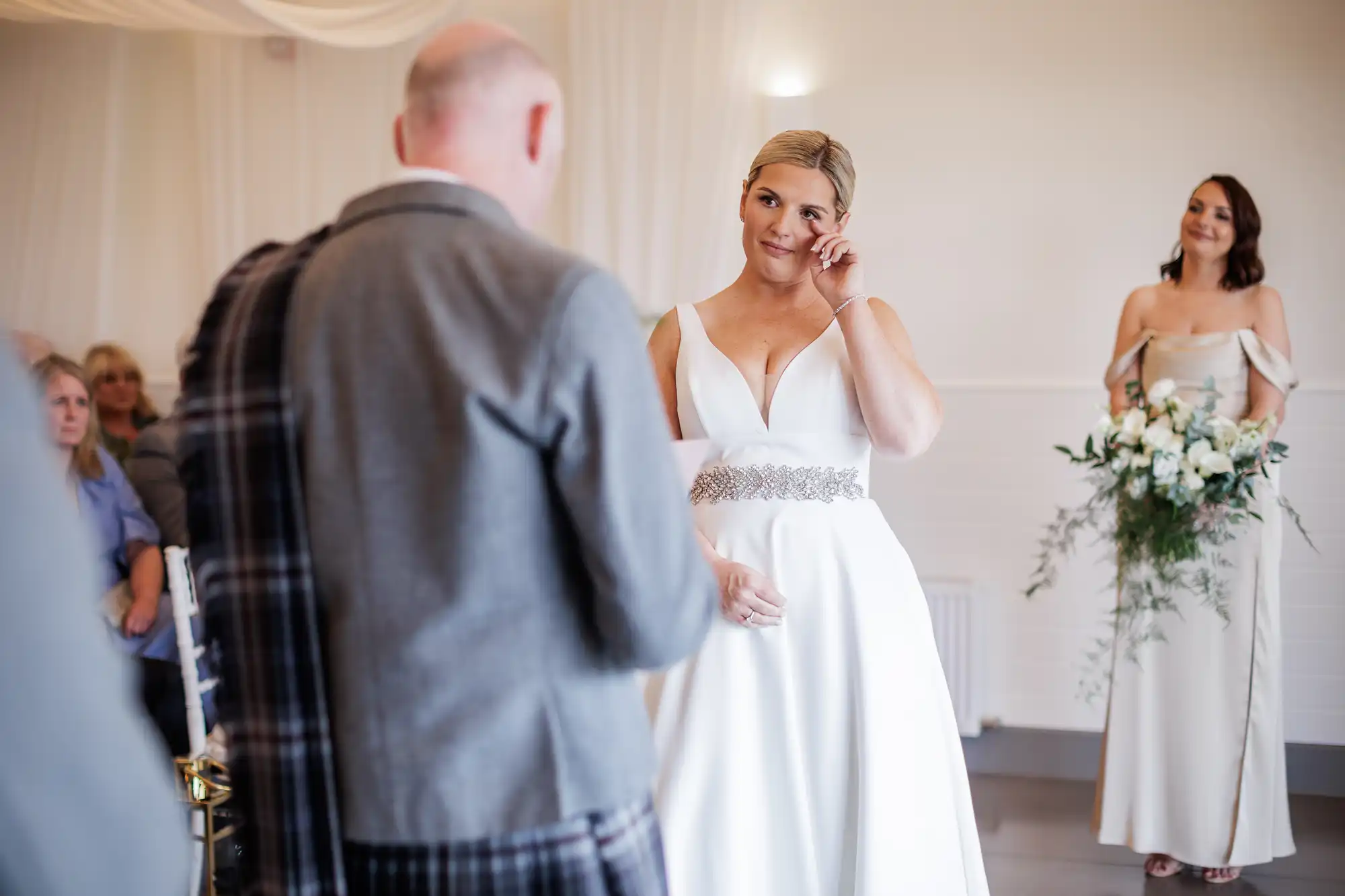 A bride in a white dress with a jewelled belt wipes a tear during a wedding ceremony. She faces a man in a grey suit with a tartan sash. A bridesmaid in a light dress stands behind, holding a bouquet. Seated guests watch in the softly-lit room.