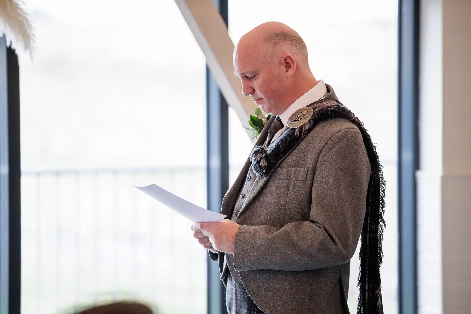 A bald man in a grey suit with a ceremonial sash and brooch reads from a sheet of paper. He stands indoors by large windows with a focused expression, holding the paper with both hands. A green buttonhole is pinned to his lapel.