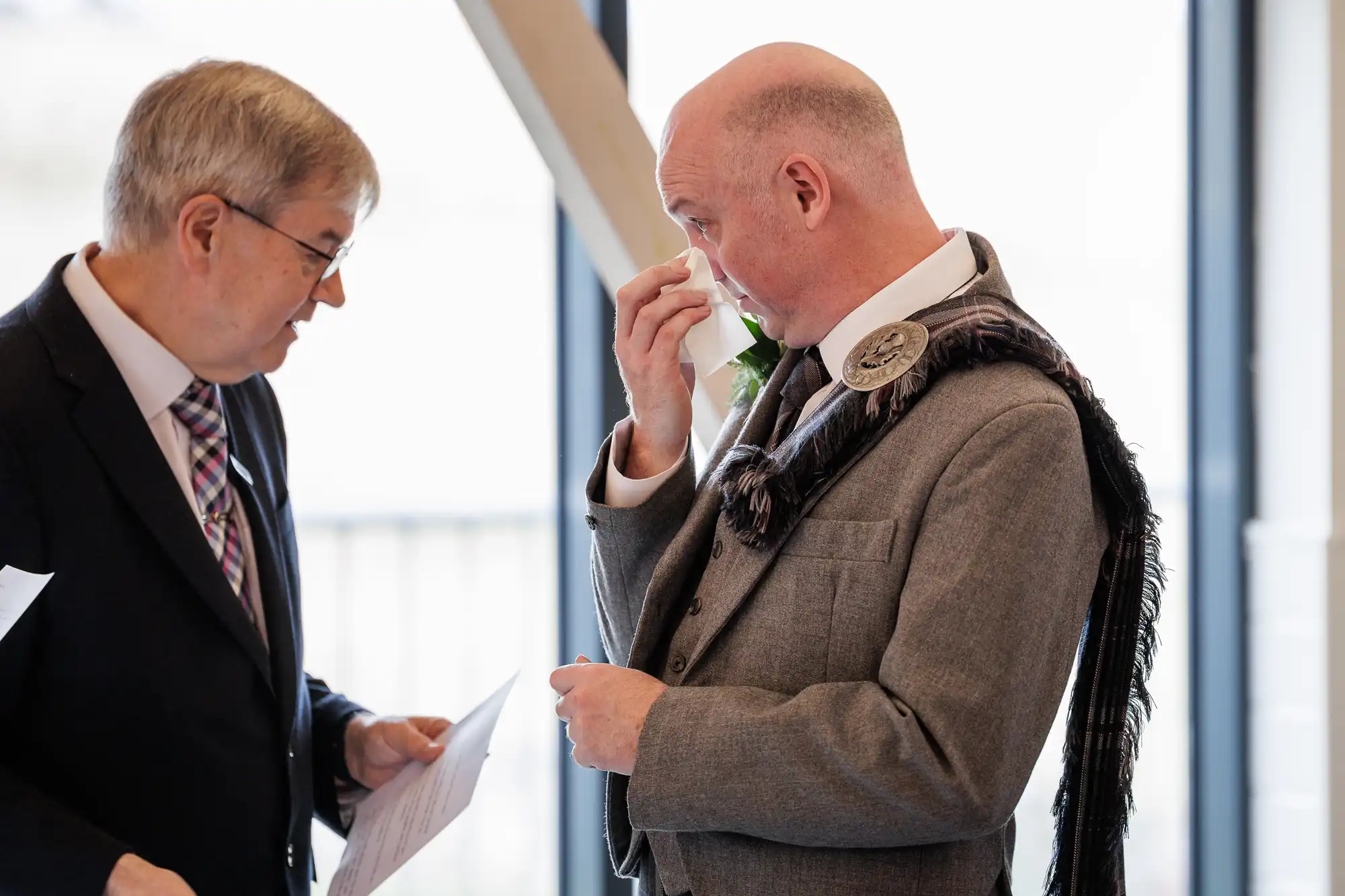 Two men dressed formally stand indoors. One, holding papers, speaks to the other, who is wearing a grey suit with a tartan sash and wiping his eye emotionally, possibly crying. The background is softly lit and out of focus.