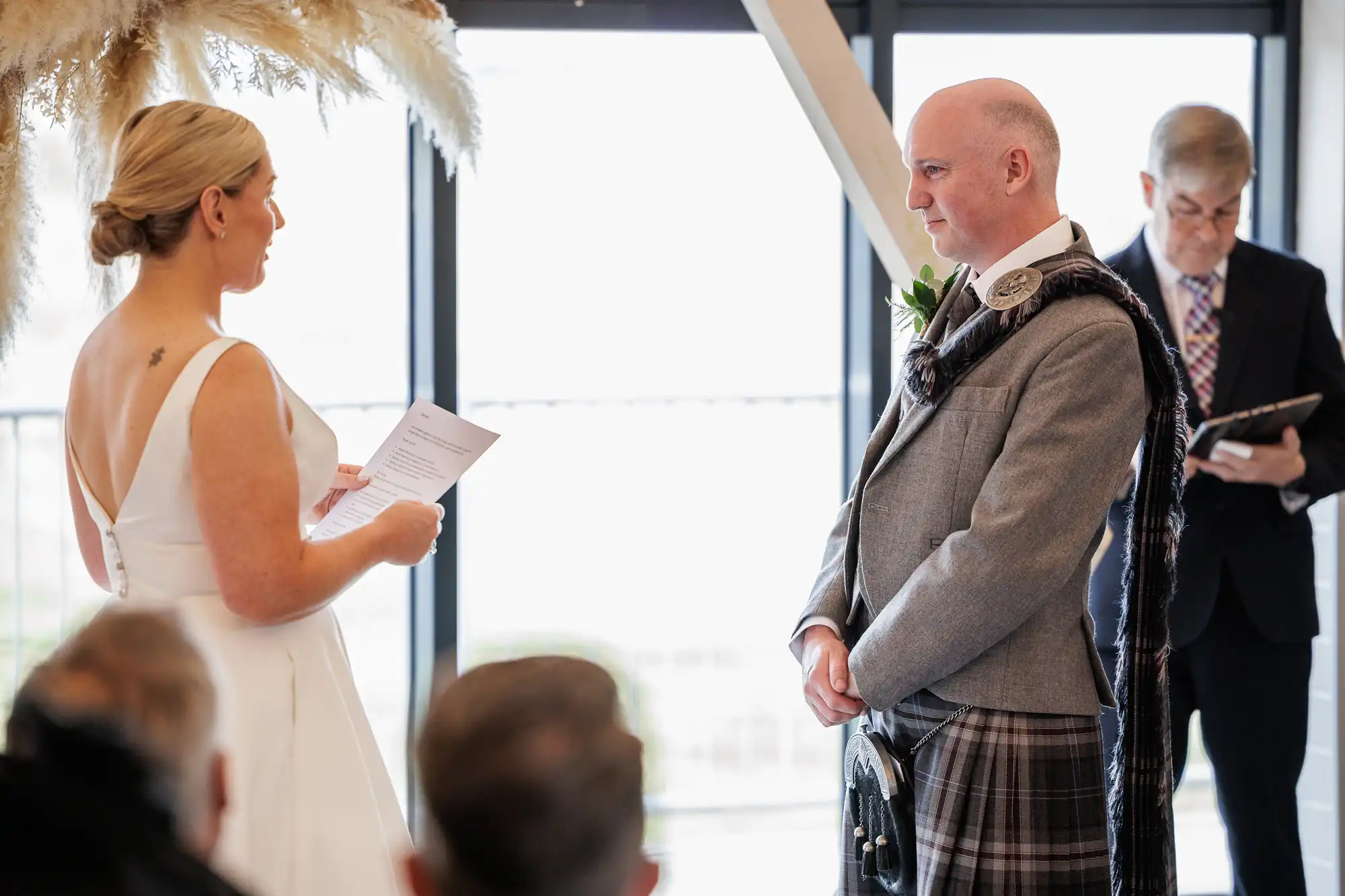 A bride in a white dress reads vows to a groom in a kilt and grey jacket during a wedding ceremony. They face each other, smiling, with guests seated in the foreground and an officiant standing behind reading from a tablet.