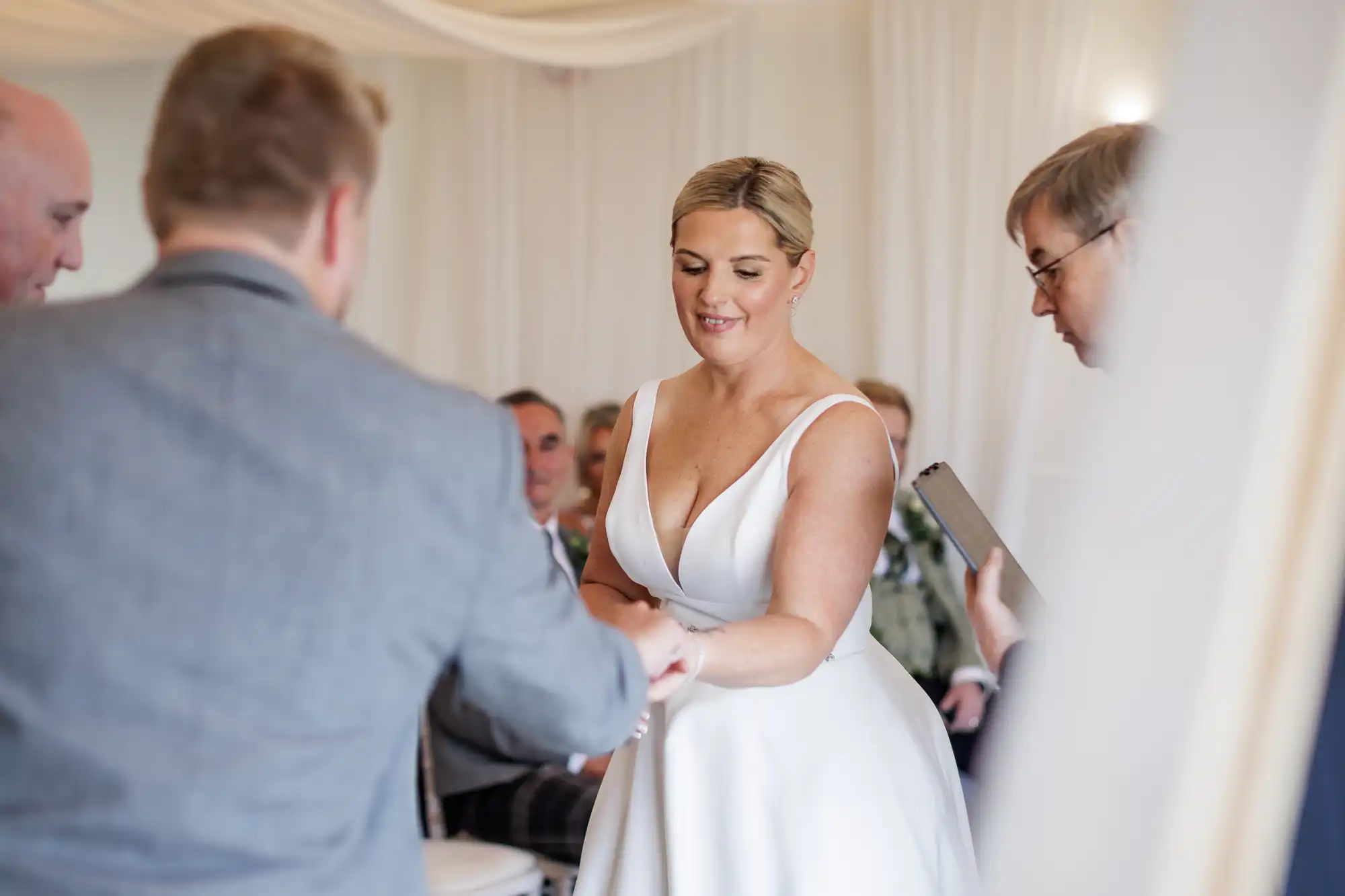 A bride in a white dress smiles softly as she holds hands with her partner during a wedding ceremony. Two men, one in a suit and one holding a folder, officiate. The setting is bright and decorated in white, with seated guests visible in the background.
