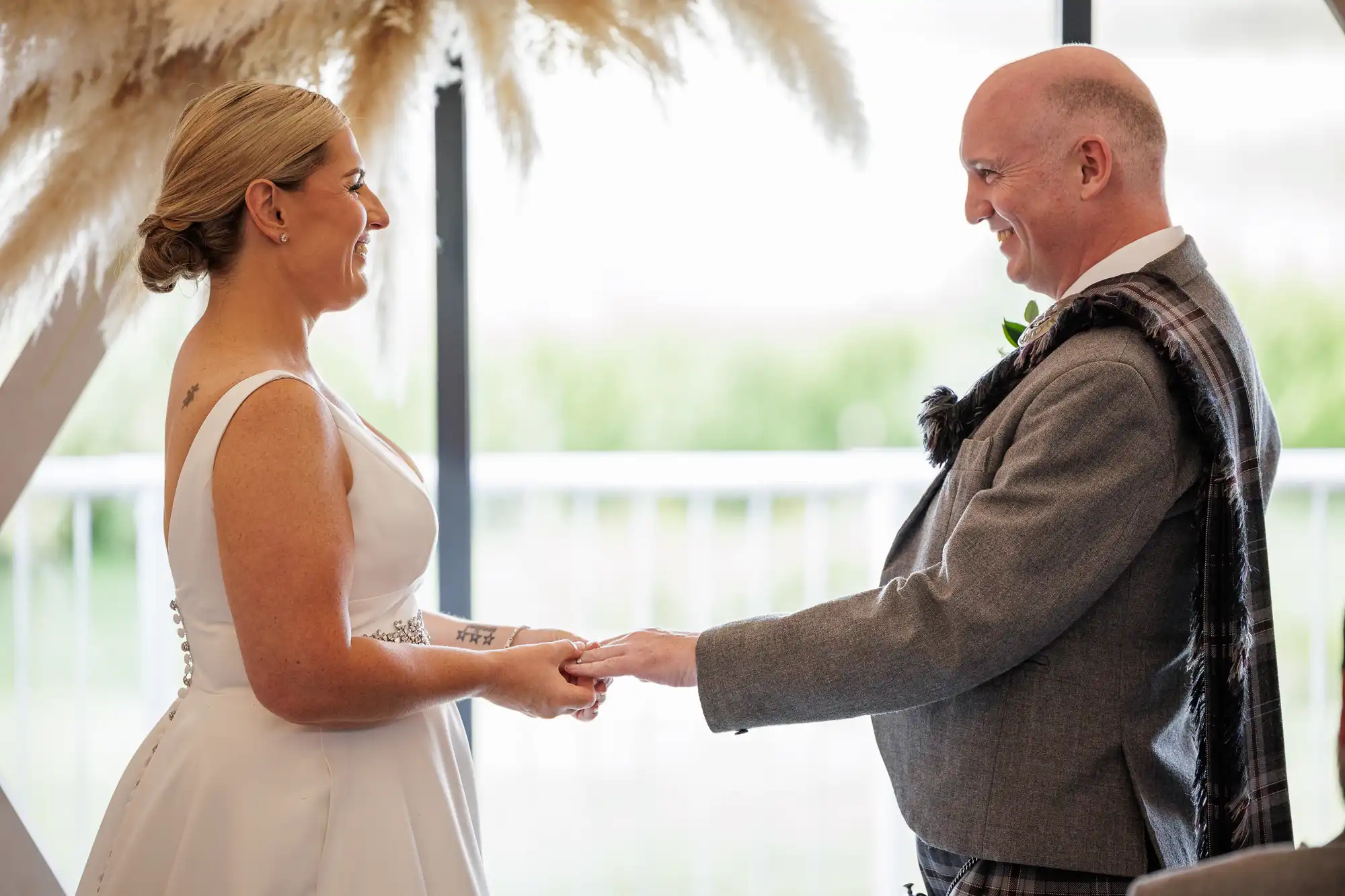 A bride in a white sleeveless dress and a groom in a grey suit with a tartan sash smile warmly at each other whilst holding hands during their wedding ceremony, standing indoors with soft natural light and pampas grass decor in the background.