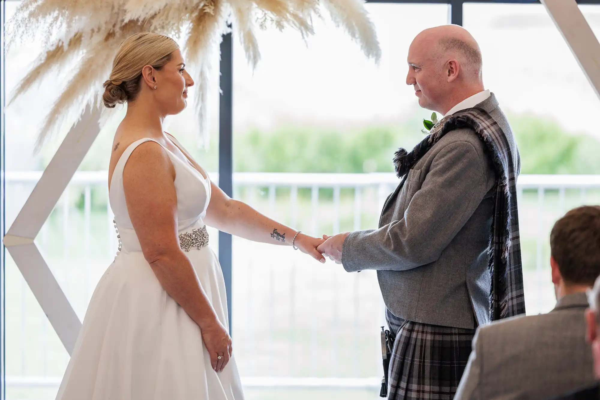 A bride in a white dress and a groom in a grey suit with a tartan sash stand facing each other, holding hands, during an indoor wedding ceremony. Decorative pampas grass is behind them, and soft daylight streams through large windows.