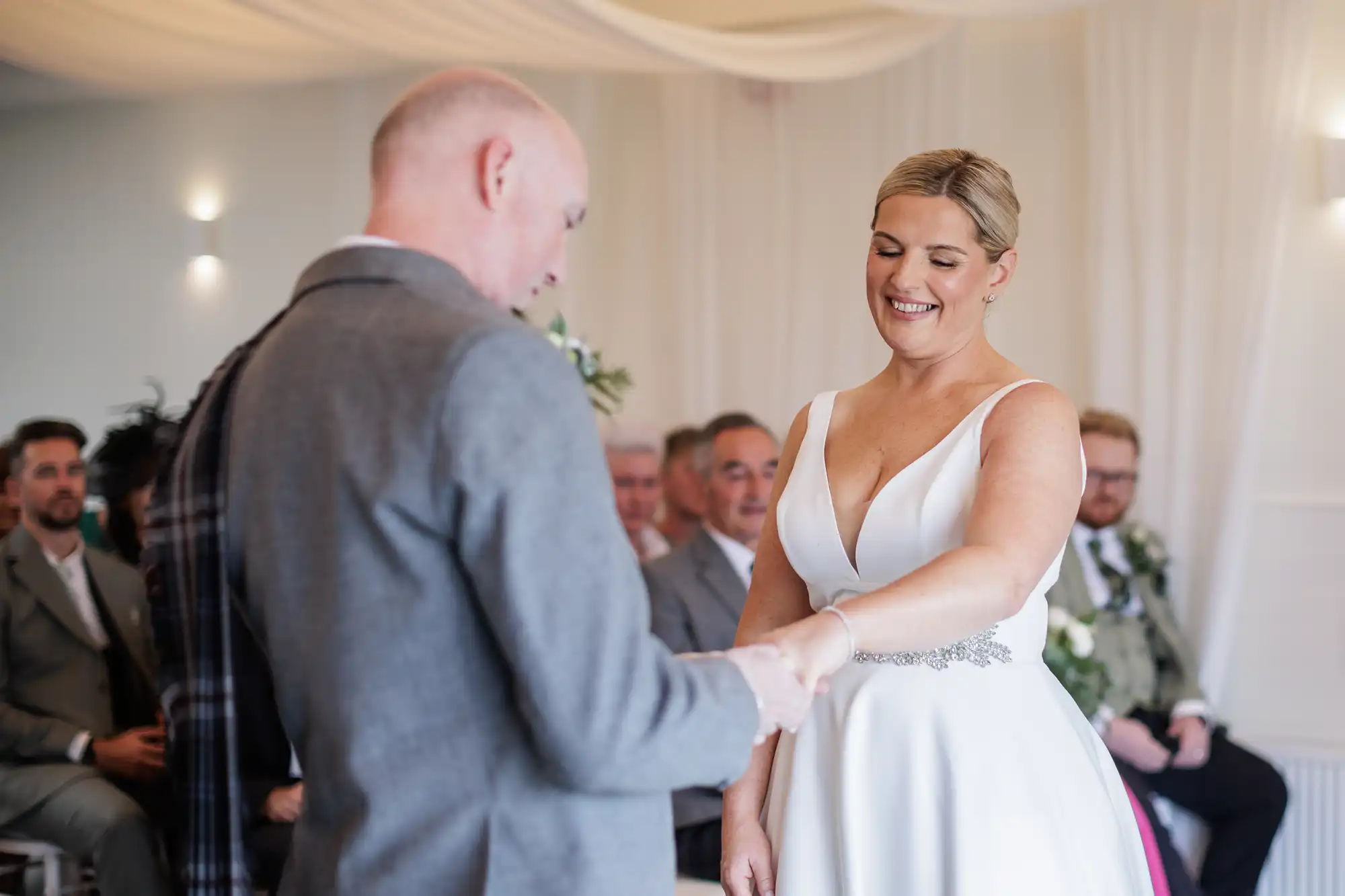 A bride in a white dress smiles as she holds hands with a groom in a grey suit during a wedding ceremony. Guests in formal attire sit in the background, watching the couple. The setting is a softly lit indoor venue with white draped fabric overhead.