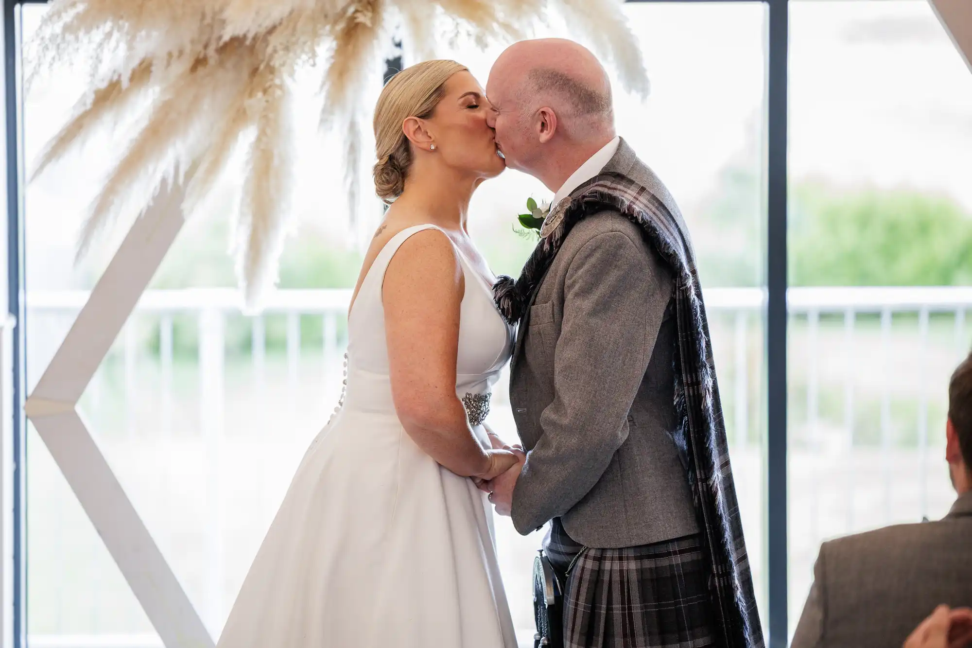 A bride in a white gown and a groom in a grey jacket with a tartan sash kiss at their wedding ceremony, holding hands. Dried pampas grass decorates the background, and a window lets in natural light behind them.
