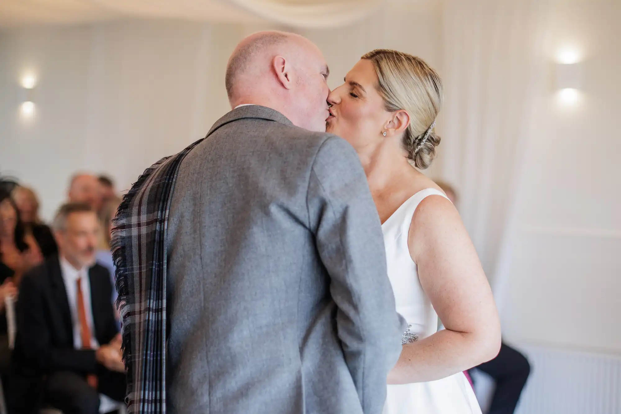 A bride and groom kiss at their wedding ceremony. The groom wears a grey suit with a tartan sash, and the bride is in a white dress. Out-of-focus guests are seated in the background, and the room is decorated in white with soft lighting.