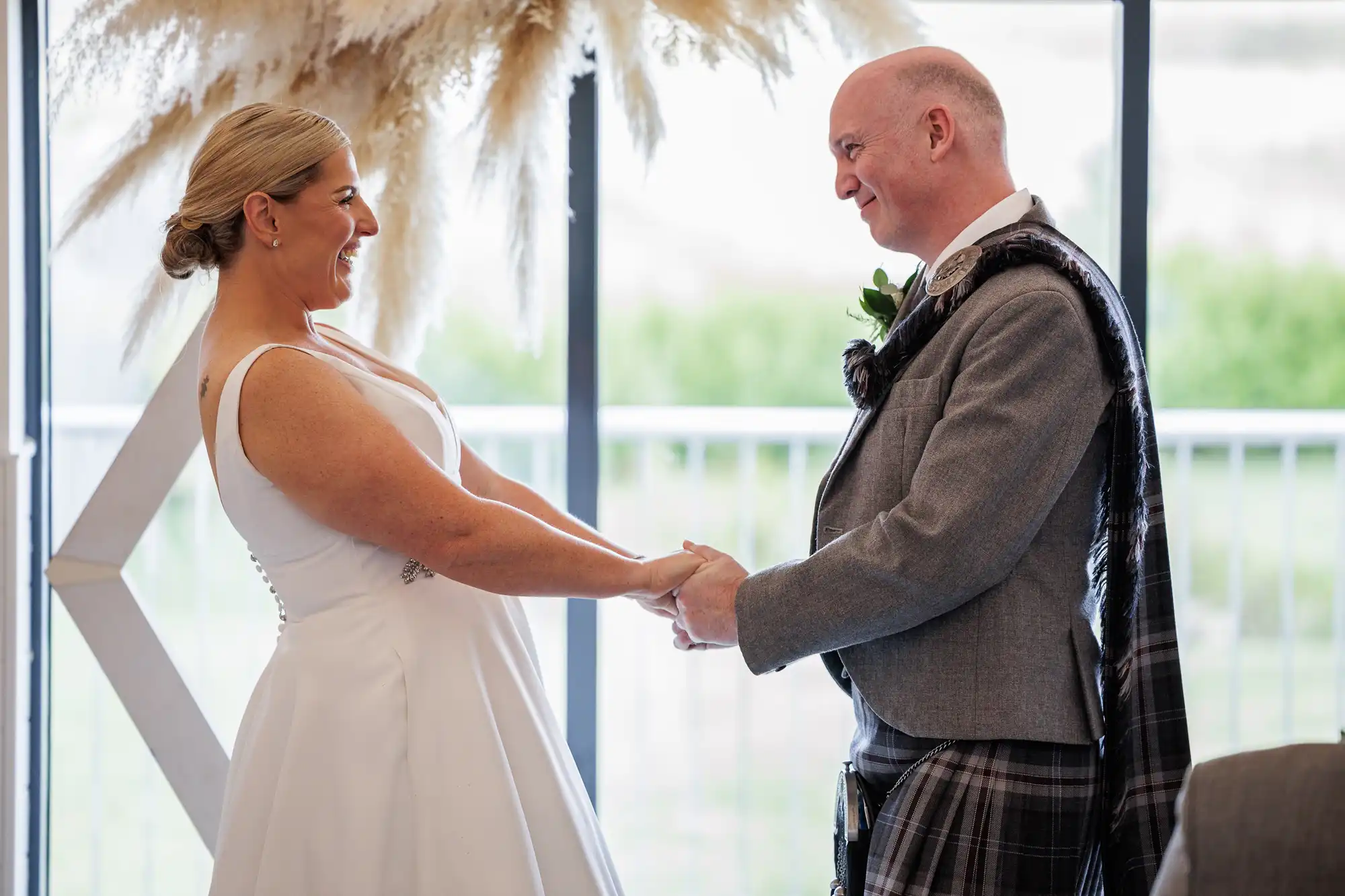 A bride in a white sleeveless gown and a groom in a grey jacket with a tartan sash hold hands and smile at each other indoors, standing near a large window with natural light and pampas grass decor in the background.