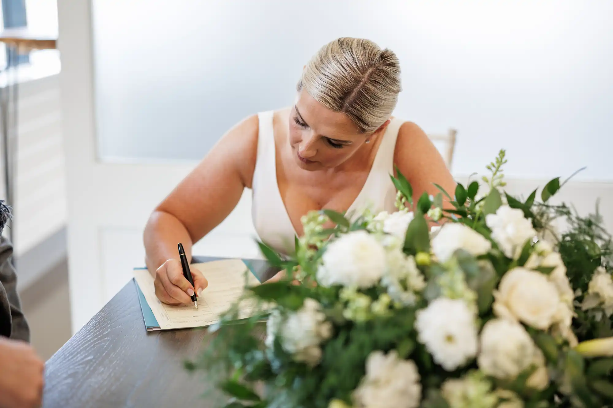 A woman with blonde hair in a white sleeveless dress sits at a table, signing a document with a pen. In front of her is a large bouquet of white flowers and green foliage. The background is softly lit with frosted glass and neutral décor.