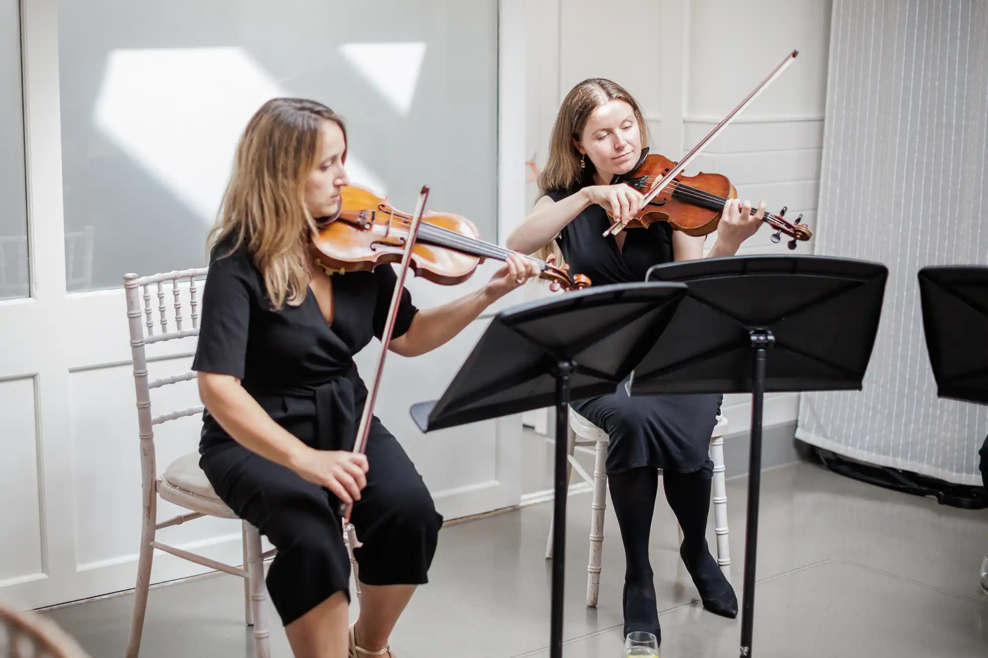 Two women in black dresses sit on chairs indoors, playing violins with sheet music on stands in front of them. Sunlight streams in from a window behind, creating bright shapes on the white wall. The mood is focused and calm.