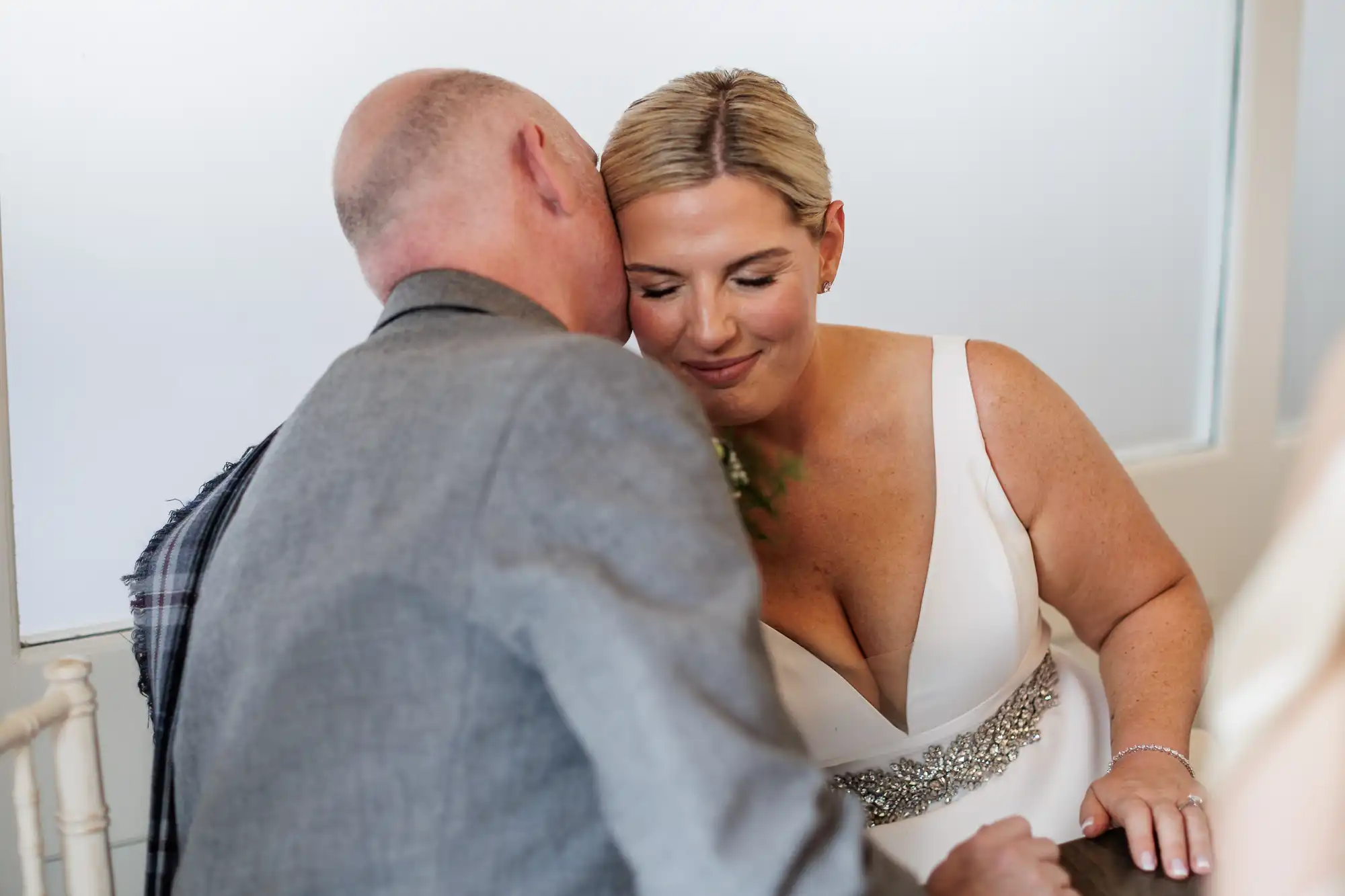 A bride in a white dress with a jewelled belt smiles gently with closed eyes as a man in a grey suit leans in close, whispering to her. They appear to share an intimate, tender moment during a wedding ceremony.