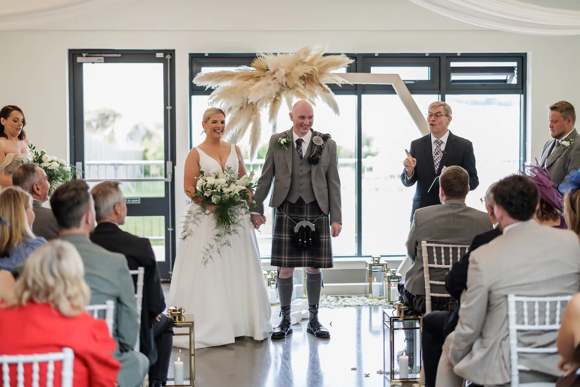 A bride in a white gown and a groom in a grey jacket and kilt stand smiling at the altar. Guests face them. Behind the couple is a modern arch with pampas grass decor. A celebrant gestures whilst speaking. Large windows let in natural light.