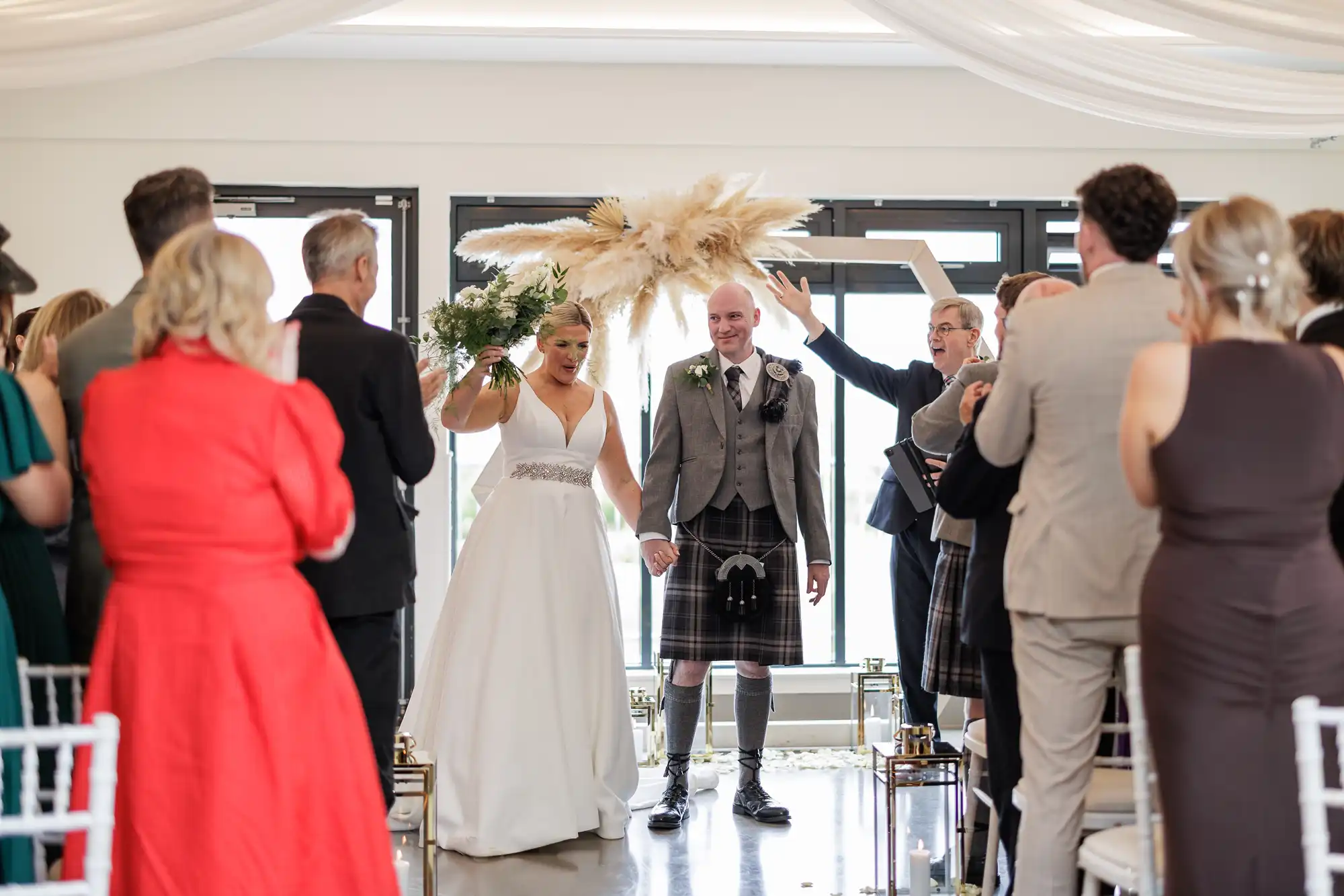 A smiling bride in a white dress and groom in a kilt walk down the aisle holding hands as guests stand and clap. The bride raises a bouquet overhead. Pampas grass decorates the arch behind them in a bright, modern venue.