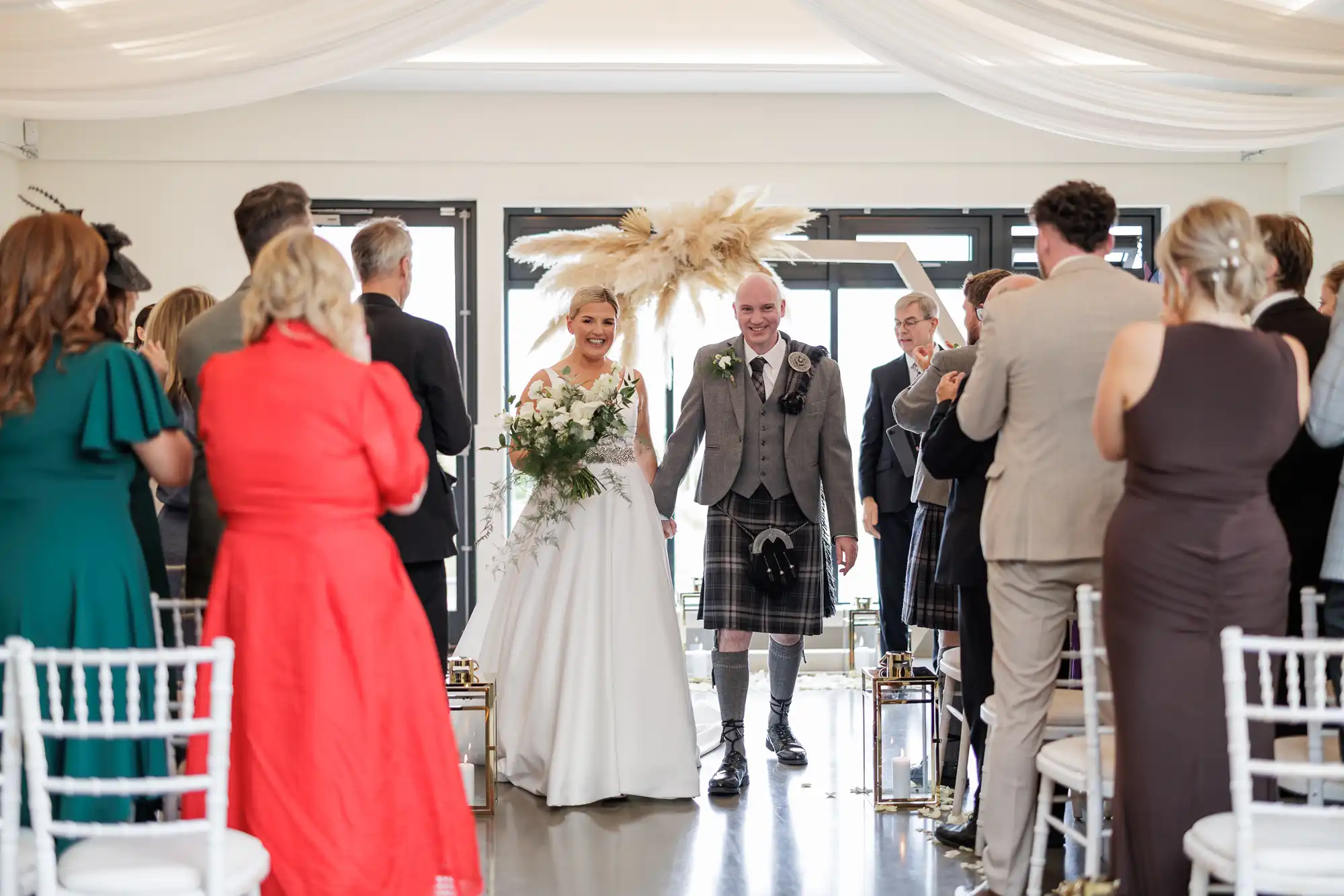 A smiling bride in a white dress and groom in a kilt walk arm in arm down the aisle, surrounded by applauding guests at an indoor wedding ceremony. Pampas grass decorates the background; people are dressed in formal attire.