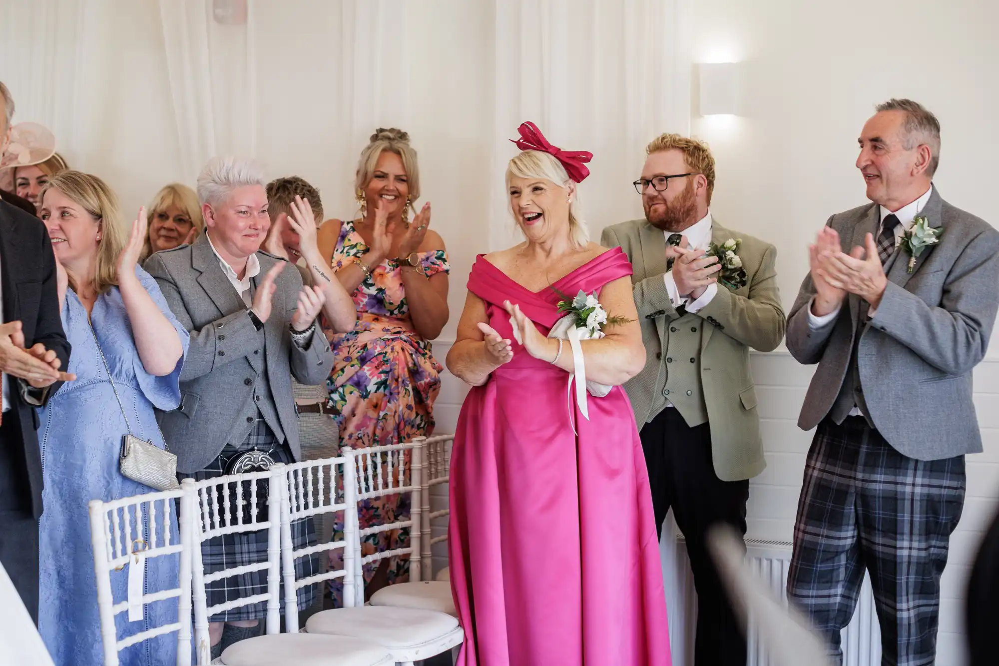 A group of people in formal attire stand and clap in a bright, white room. A woman in a bright pink dress with a bow and corsage smiles at the centre, surrounded by people in suits and floral dresses, celebrating a joyful event. White chairs are in the foreground.