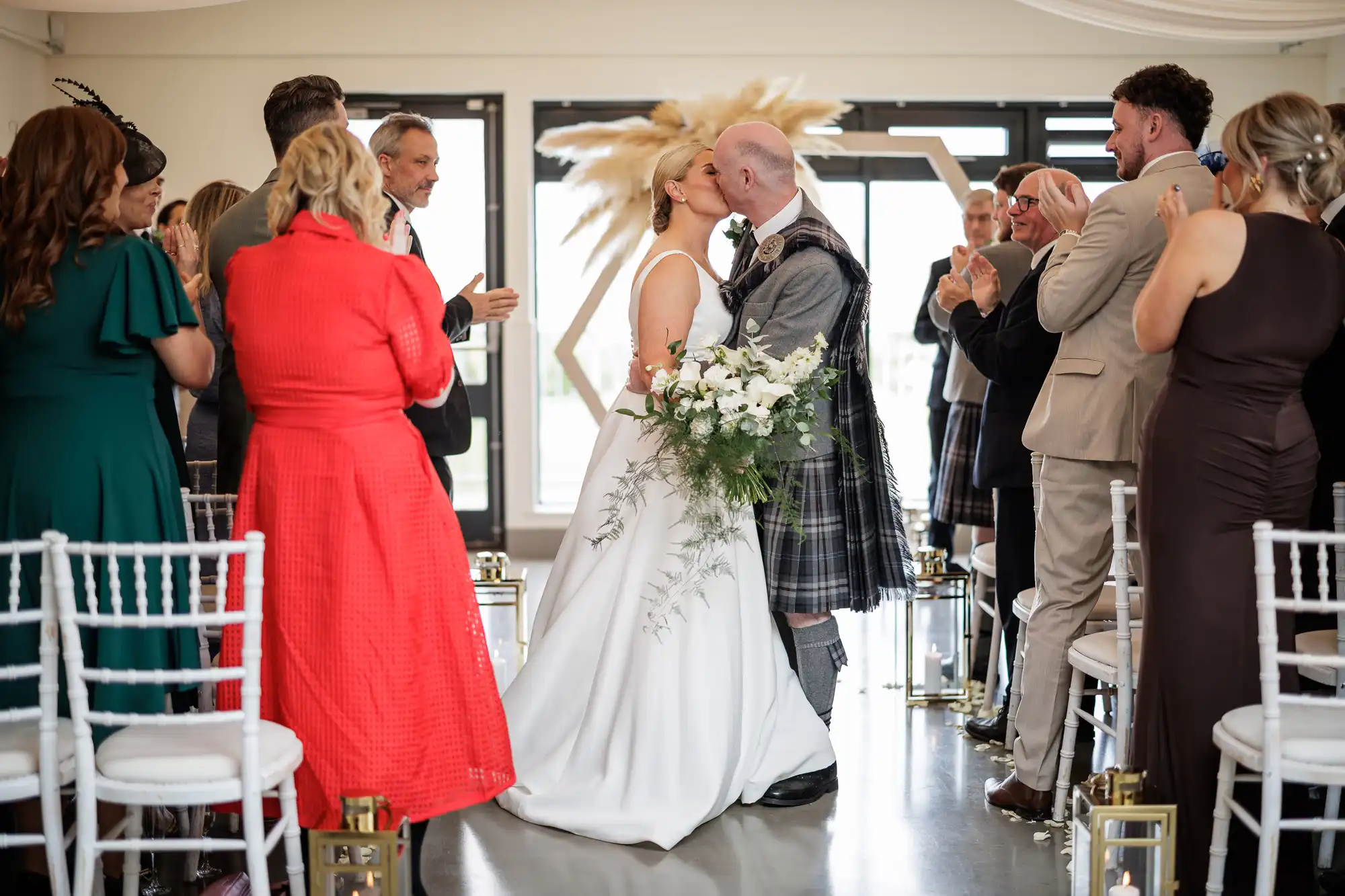 A bride in a white dress and a groom in a kilt kiss at the end of an aisle, surrounded by guests standing and clapping in a bright indoor wedding venue decorated with white chairs and floral arrangements.