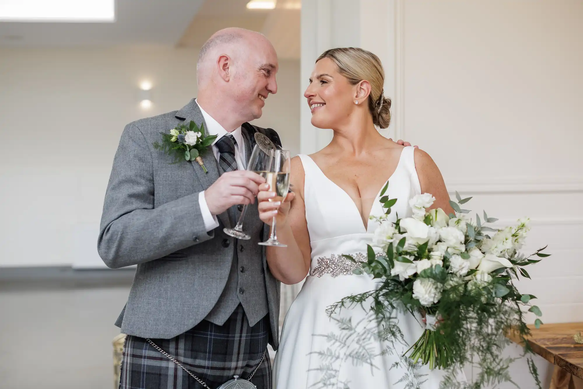 A smiling couple in wedding attire clink champagne glasses. The man wears a grey jacket, white shirt, kilt, and buttonhole. The woman wears a white dress, jewelled belt, and holds a lush bouquet of white flowers and greenery. They gaze at each other happily.