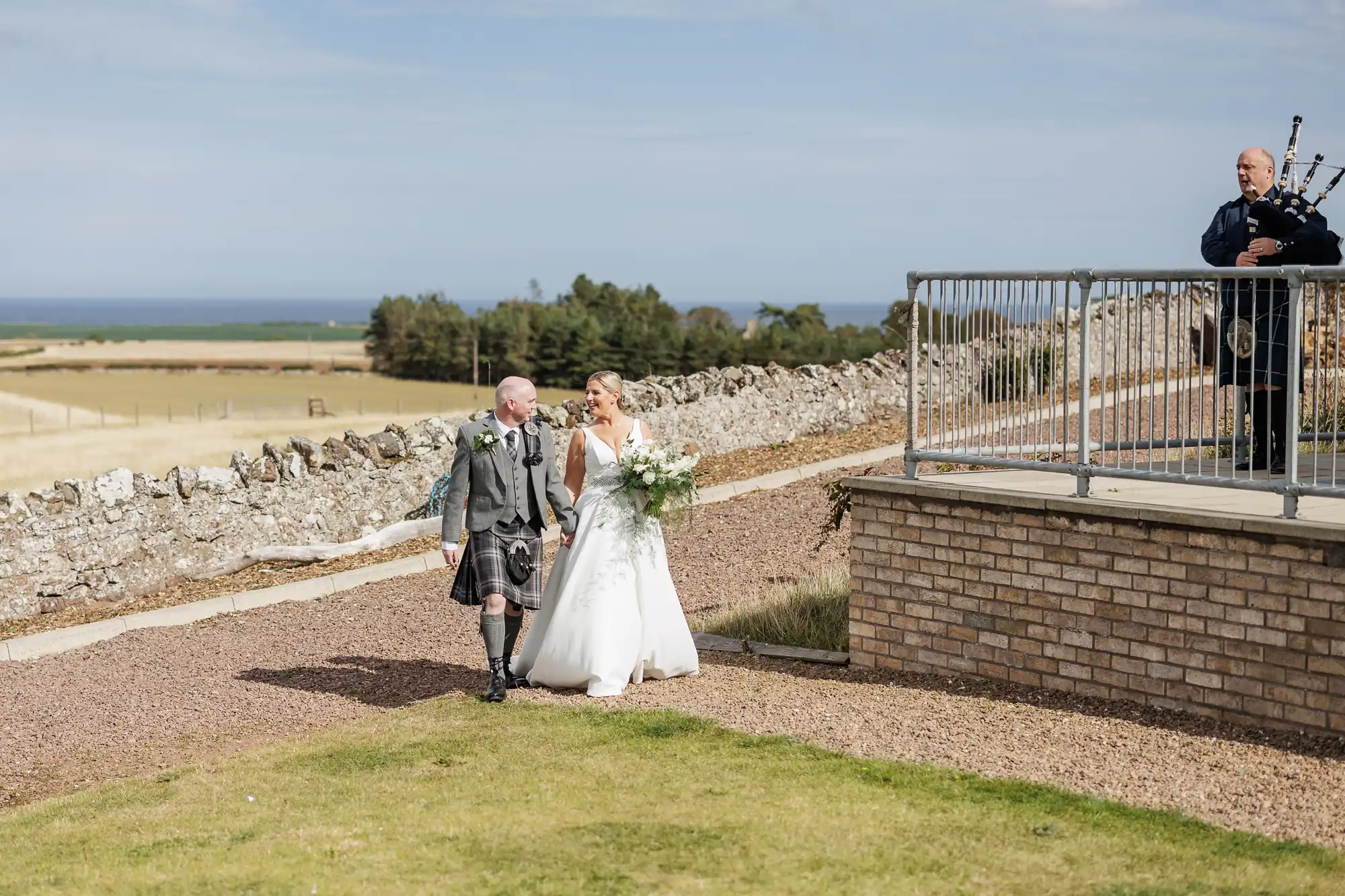 A bride in a white dress walks outside on a path, holding a bouquet and the arm of an older man in a kilt. They smile at each other. A bagpiper in traditional attire stands on a raised patio. Behind them are fields, stone walls, and a distant blue sea.