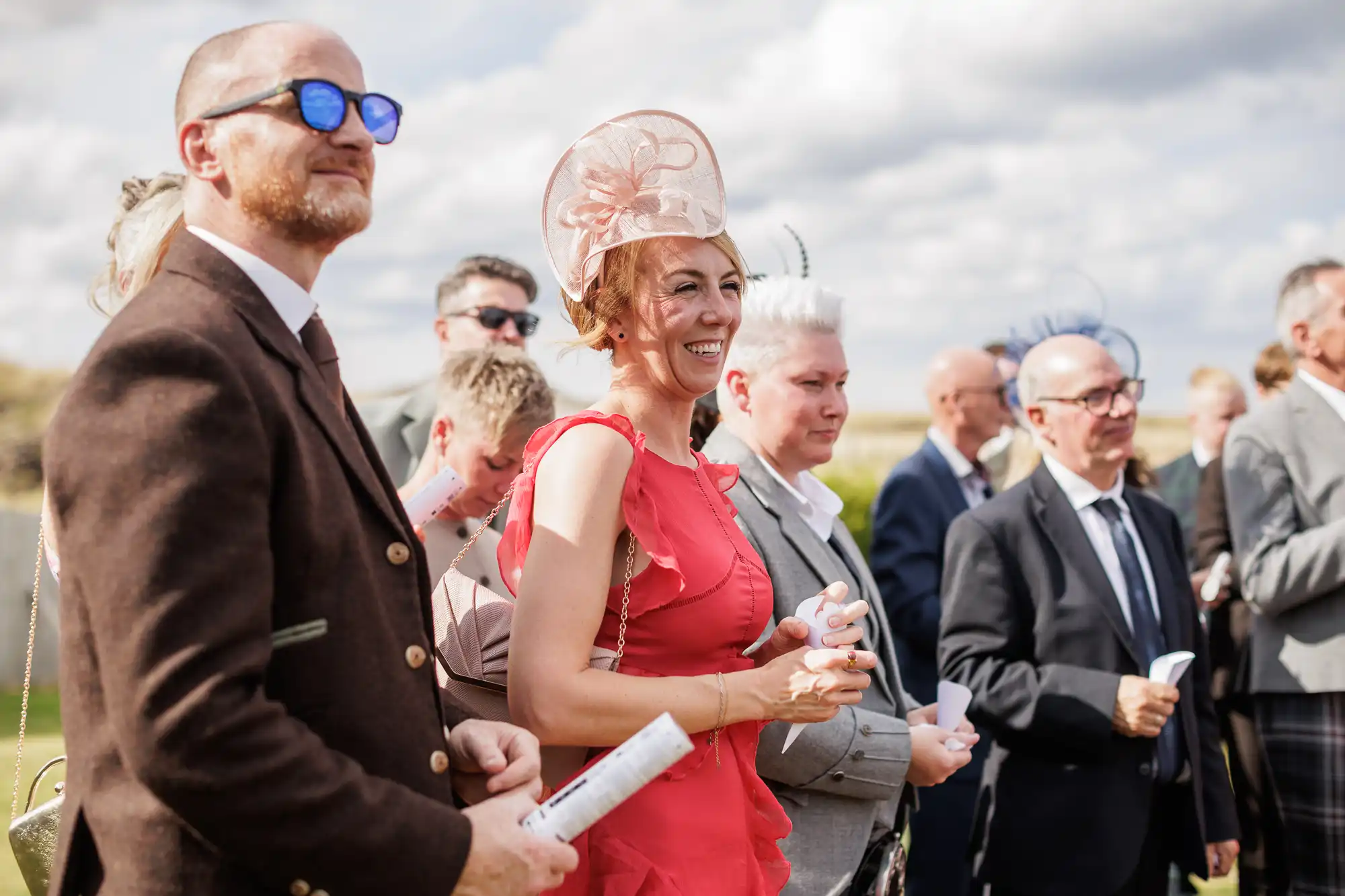 A group of people dressed formally outdoors, smiling and watching something off-camera. A woman in a bright coral dress and pink hat stands at the front, holding papers. Sunny weather, blue sky with clouds, and green grass in the background.