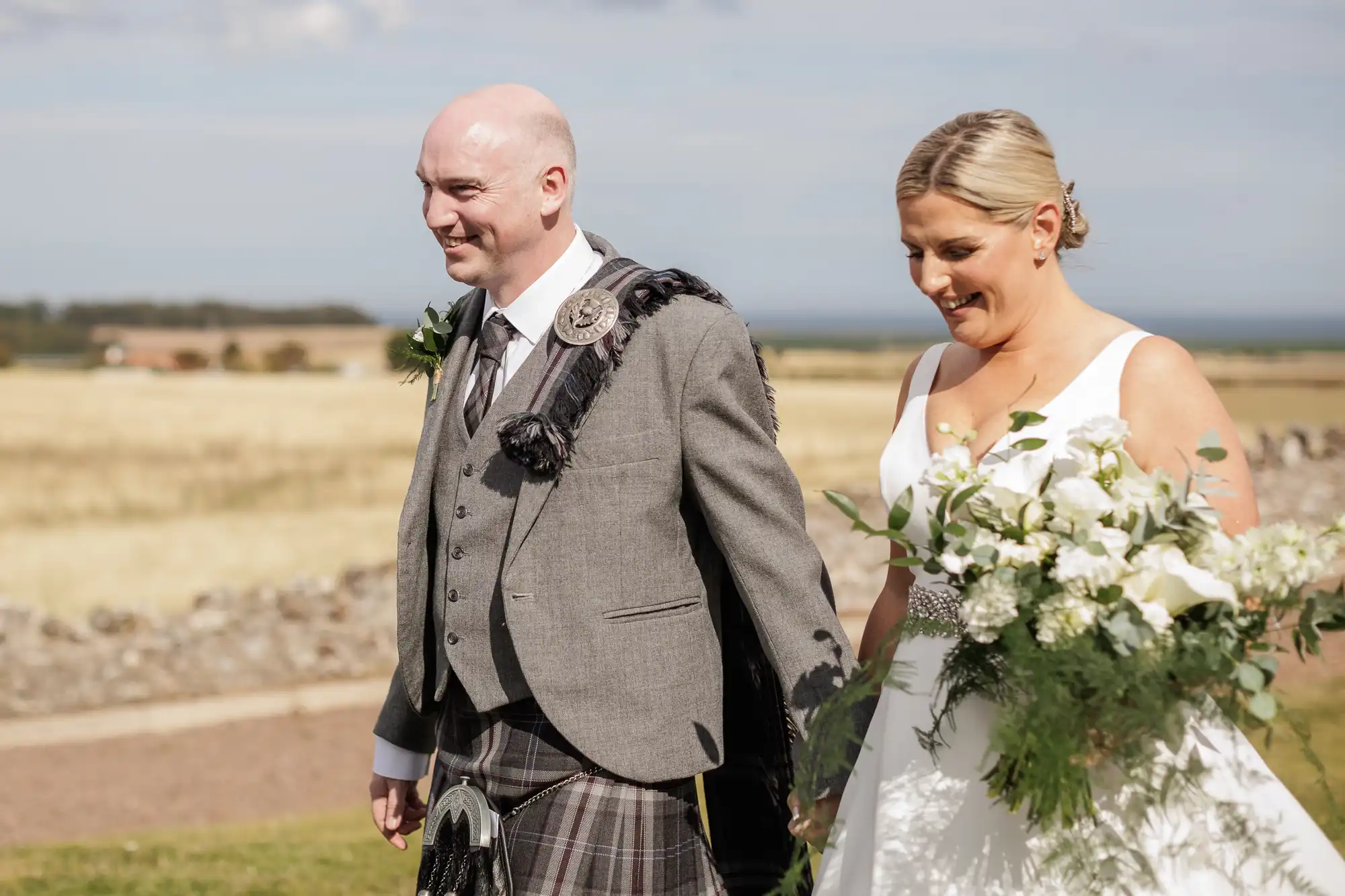 A smiling couple walks outdoors on a sunny day. The man wears a grey jacket, kilt, and plaid sash, whilst the woman wears a white sleeveless dress and holds a large bouquet of white flowers. They hold hands, with fields and a stone wall in the background.