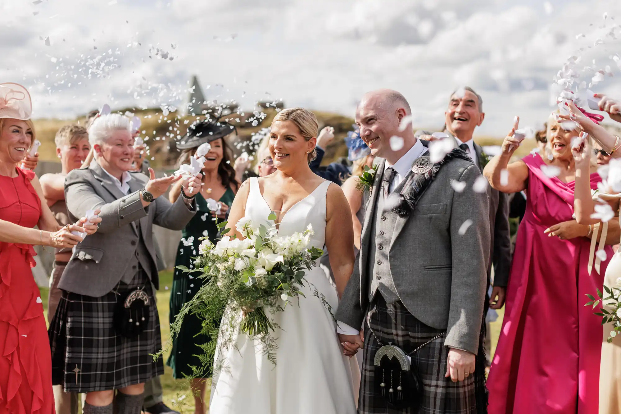 A bride in a white dress and a groom in a grey jacket and kilt smile whilst holding hands, surrounded by joyful guests in colourful outfits and kilts, tossing confetti outdoors under a partly cloudy sky. The bride holds a bouquet of white flowers and greenery.