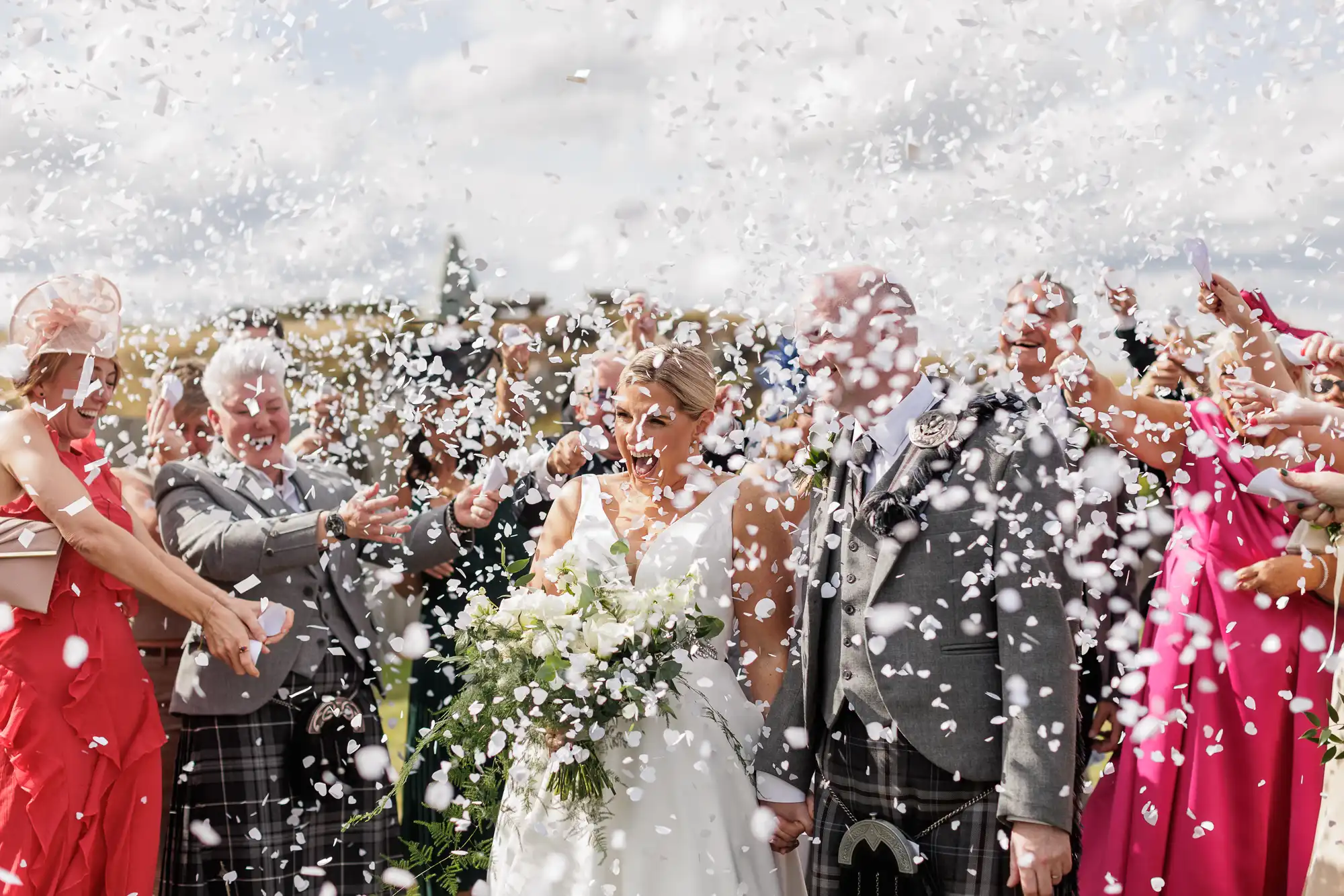A bride and groom stand smiling outdoors as guests shower them with white confetti. The bride holds a bouquet of white flowers, and both wear formal attire. Several guests in colourful outfits celebrate joyfully under a cloudy sky.