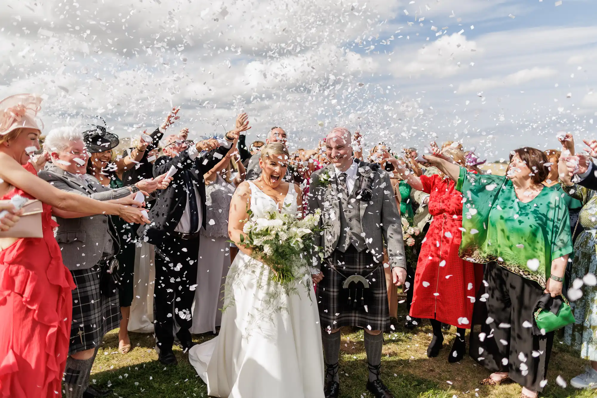 A bride in a white dress and a groom in a kilt smile as guests throw white confetti. The group, dressed in formal and colourful attire, celebrates outdoors under a partly cloudy sky. The joyful crowd surrounds the couple on green grass.