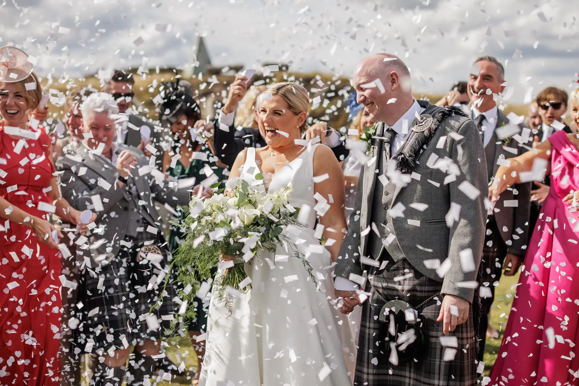 A bride in a white dress and groom in a traditional Scottish kilt smile, holding hands, surrounded by guests throwing white confetti outside. The joyful crowd wears colourful outfits and the background shows a bright, partly cloudy sky.