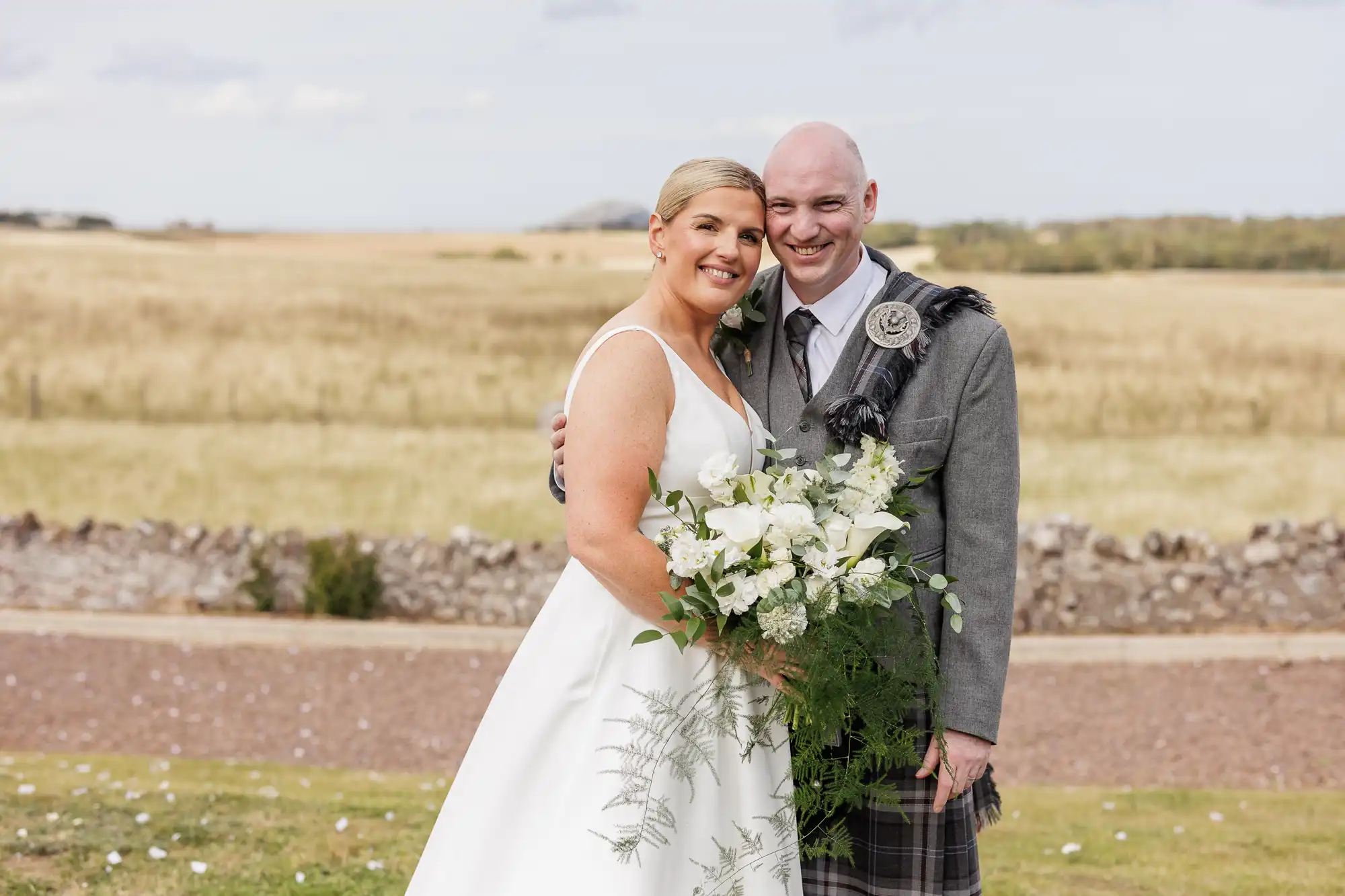 A smiling bride in a white dress holds a bouquet of white flowers while standing arm in arm with a groom in a grey jacket and tartan kilt. They are outdoors with a stone wall, grassy field, and cloudy sky in the background.