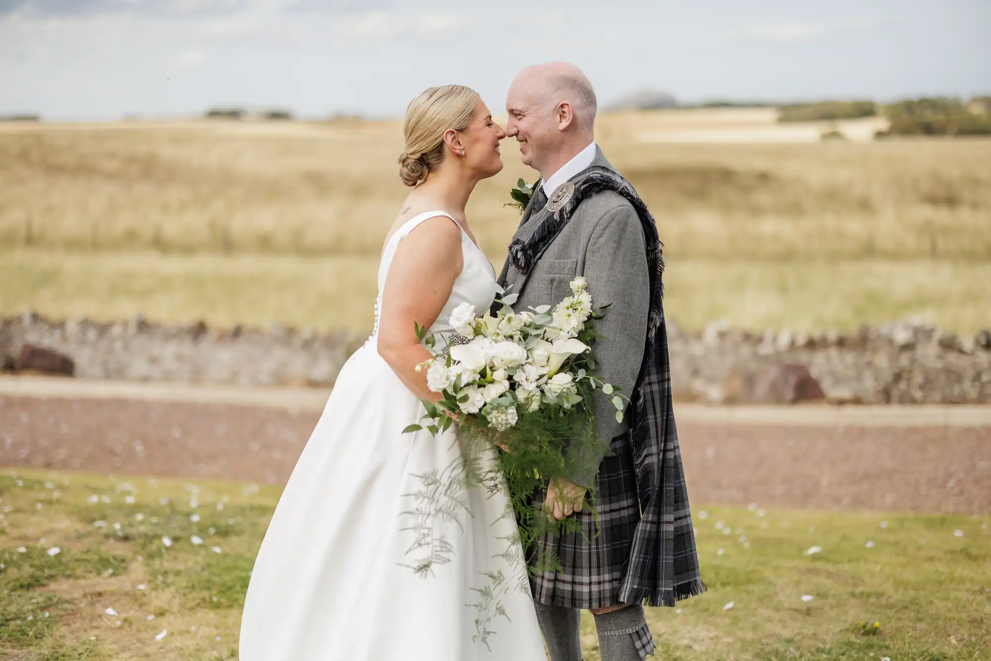 A bride in a white dress holds a bouquet of white flowers and greenery, smiling and touching noses with a groom in a grey kilt and sash. They stand outdoors on grass in front of a stone wall and an open, golden field under a cloudy sky.