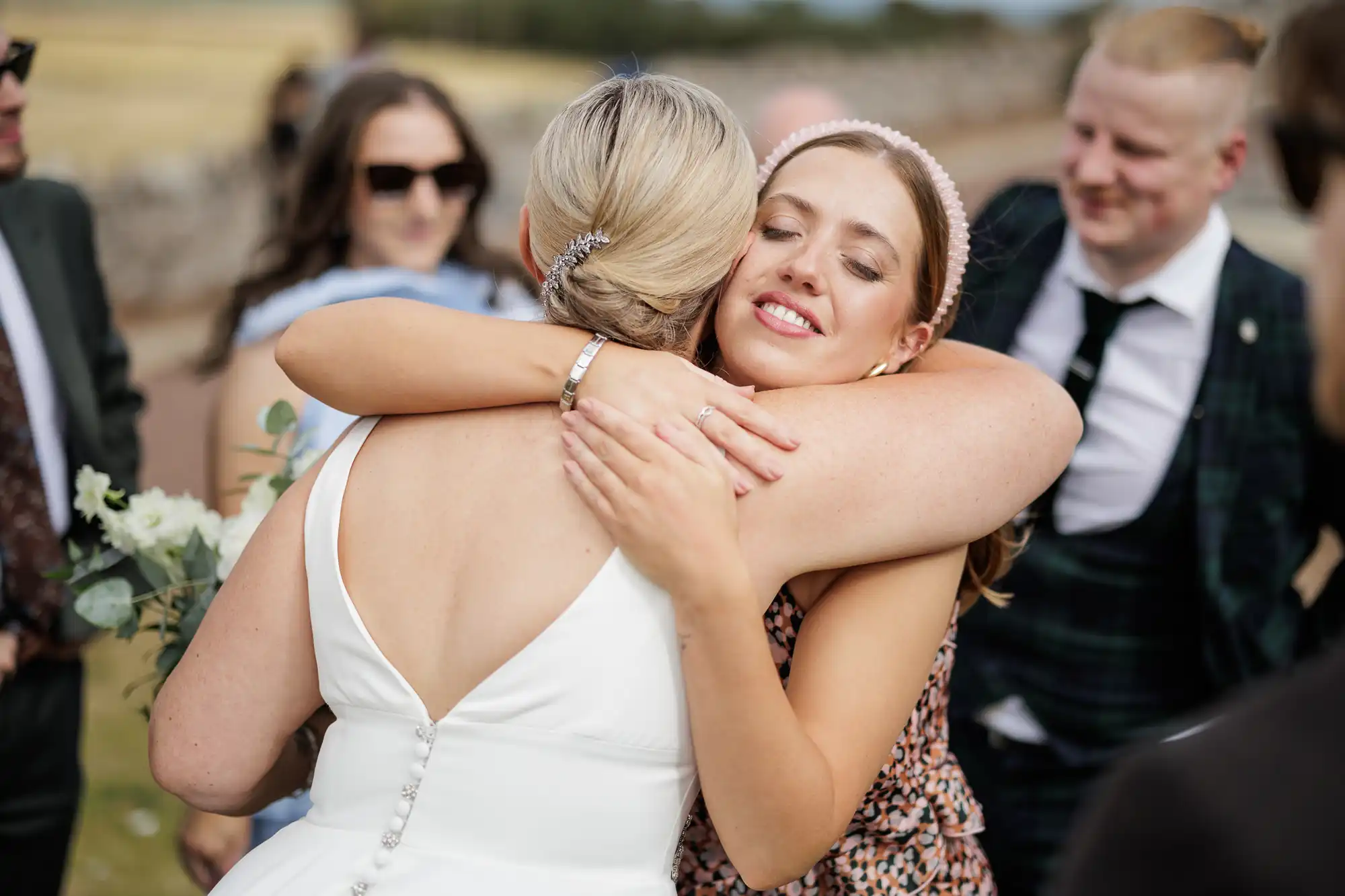 A bride in a white dress hugs a smiling woman with light brown hair and a pink headband. Other guests, dressed formally, stand nearby outdoors on a sunny day, some wearing sunglasses. The background shows a stone wall and grassy fields.