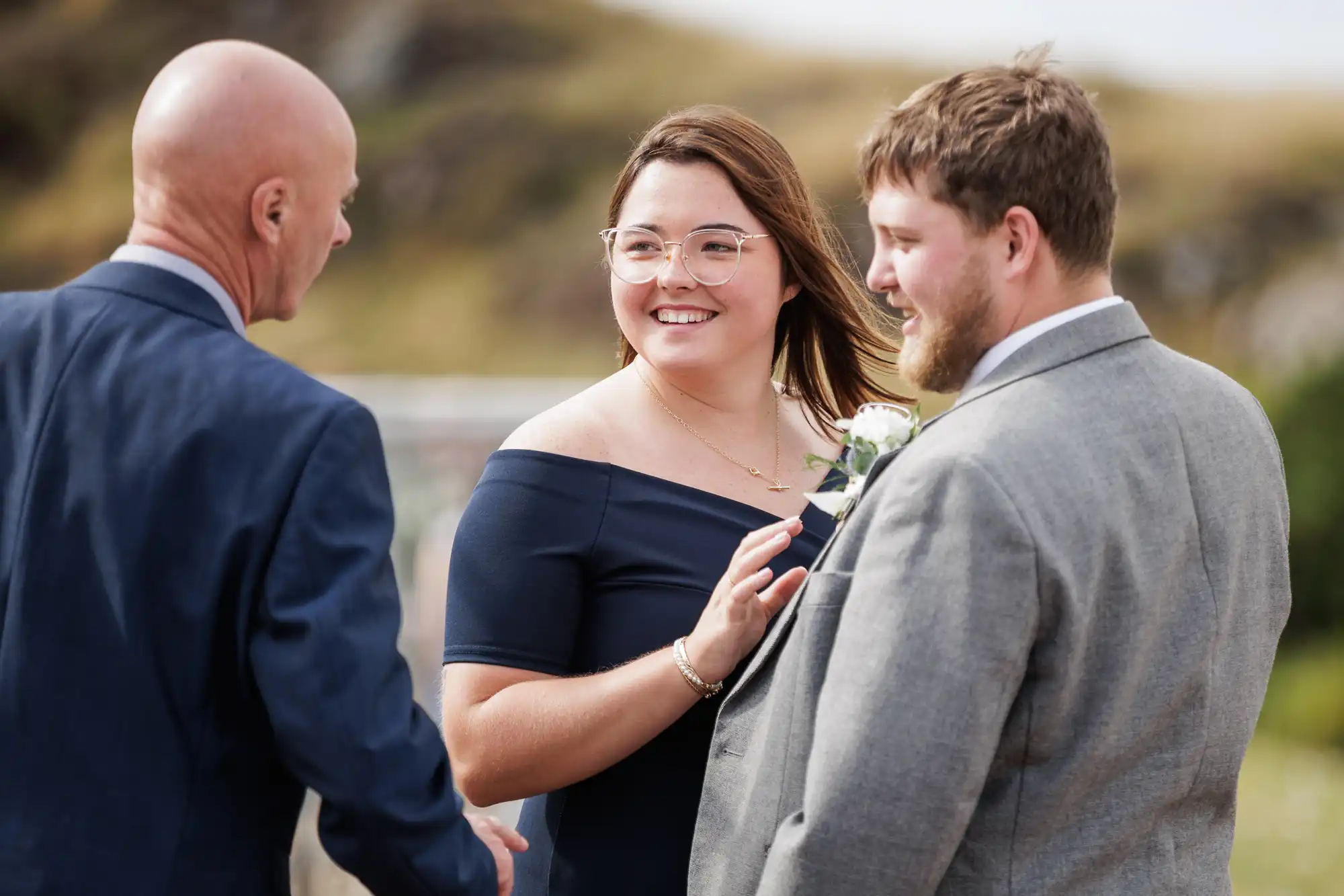 Three people stand outdoors, dressed formally. A woman in an off-shoulder navy dress smiles whilst talking to two men in suits. One man is bald, the other has a beard and buttonhole. The background is blurred greenery and hills.
