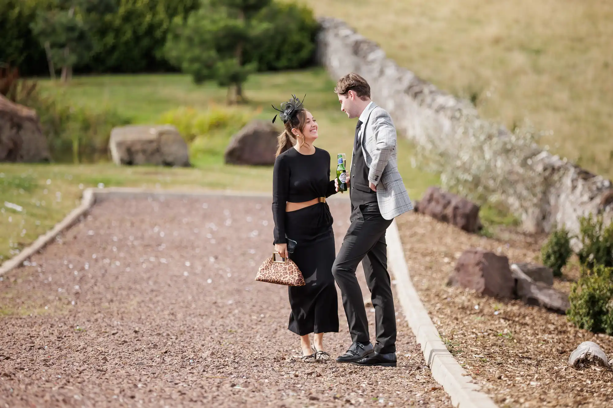 A woman in a black dress, fascinator, and animal print handbag walks beside a man in a light grey blazer and black trousers. They smile at each other on a gravel path, with green grass and large rocks in the background. The man holds a tin.