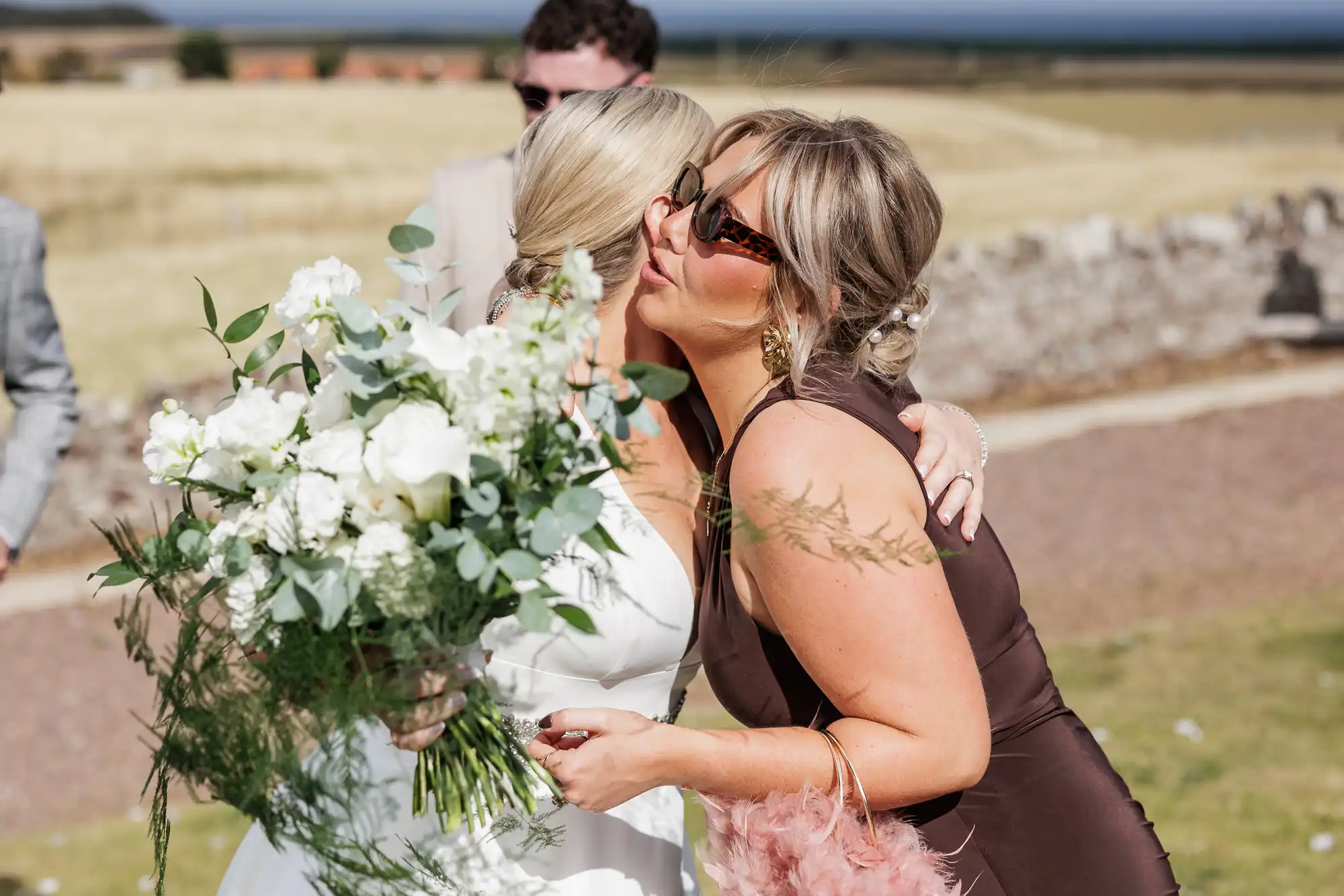 A woman in a white wedding dress holding a large white bouquet is kissed on the cheek by a woman in a brown dress and sunglasses at an outdoor event, with dry grass fields and a stone wall in the background.