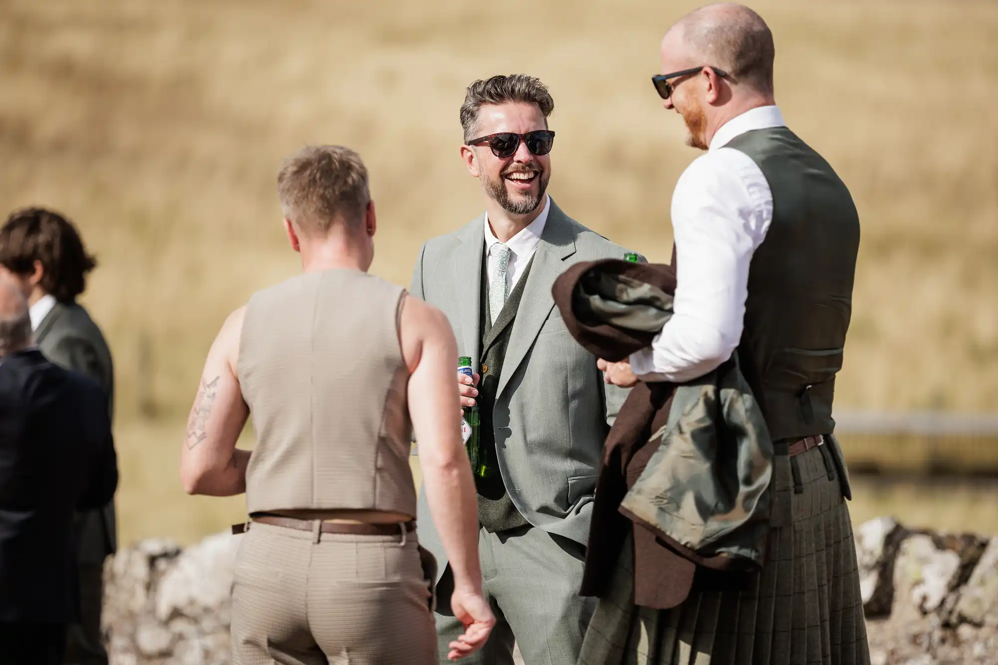 Three men stand outdoors, smiling and talking. One wears a green suit and sunglasses, holding a drink. Another has a shaved head and wears a kilt with a waistcoat. The third, seen from behind, wears a light beige outfit. The background is a grassy, sunlit field.