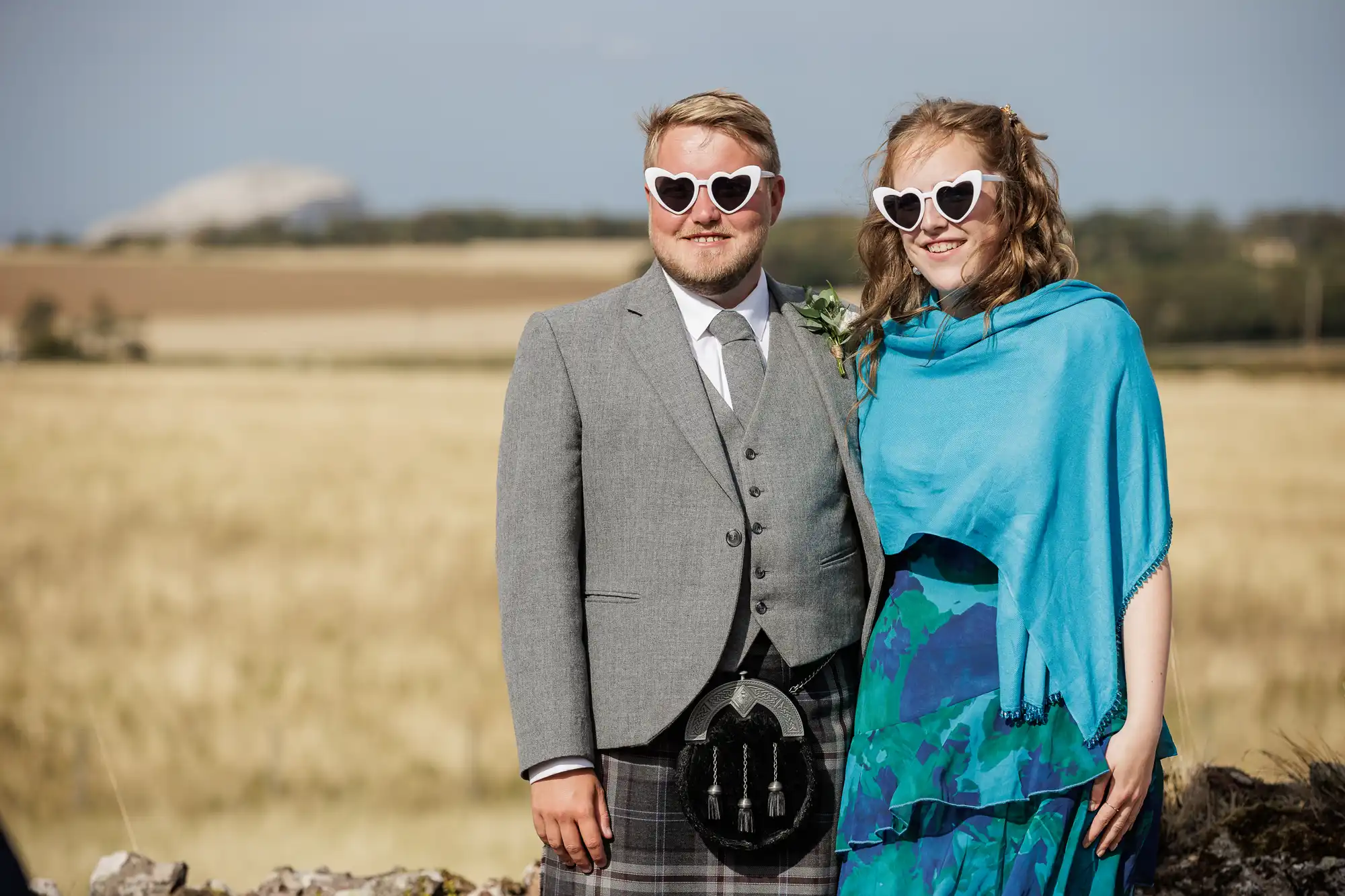 A man in a grey jacket, kilt, and tie stands next to a woman in a blue-green patterned dress with a blue shawl. Both wear white heart-shaped sunglasses and smile, posing outdoors in front of a stone wall and golden field under a blue sky.