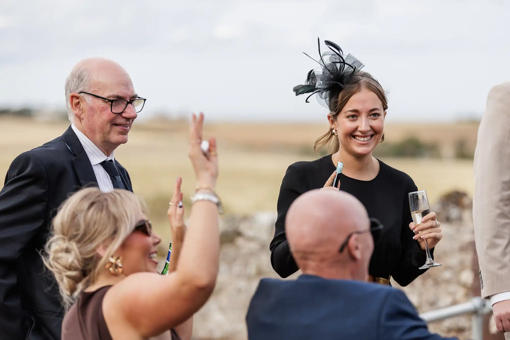 Four people at an outdoor event dressed formally. A smiling woman holds a glass of champagne and wears a black dress and fascinator. Two seated people wave, while a man in glasses and a suit stands nearby. Fields and sky are visible in the background.