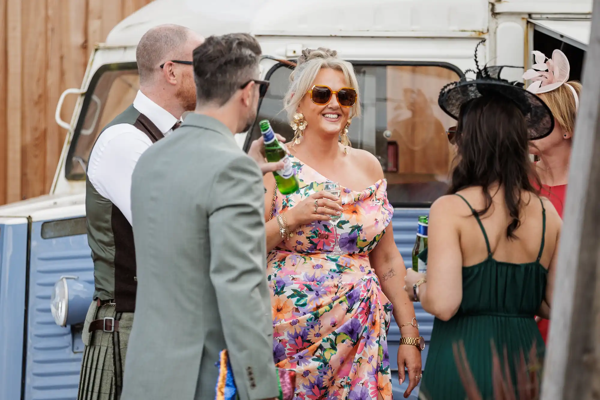 Four adults stand outside near a vintage blue vehicle, holding bottles and smiling. The central woman wears a colourful floral dress and sunglasses, while the others wear formal attire, including hats and suits, suggesting a festive outdoor event.
