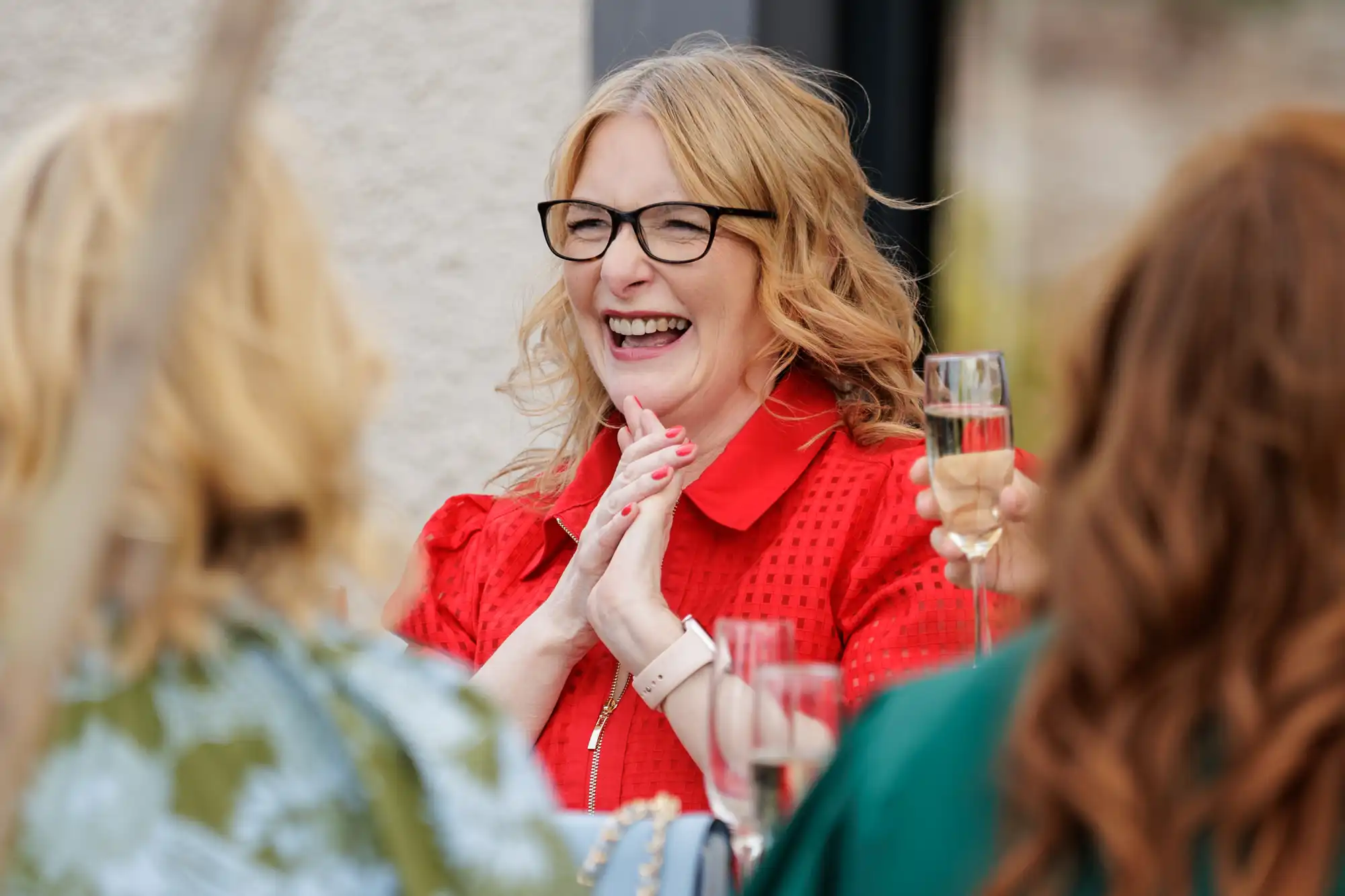 A woman with wavy blonde hair and black glasses, wearing a bright red top, laughs joyfully whilst holding her hands together. She is outdoors, surrounded by two other women and people holding champagne flutes, suggesting a celebratory gathering.